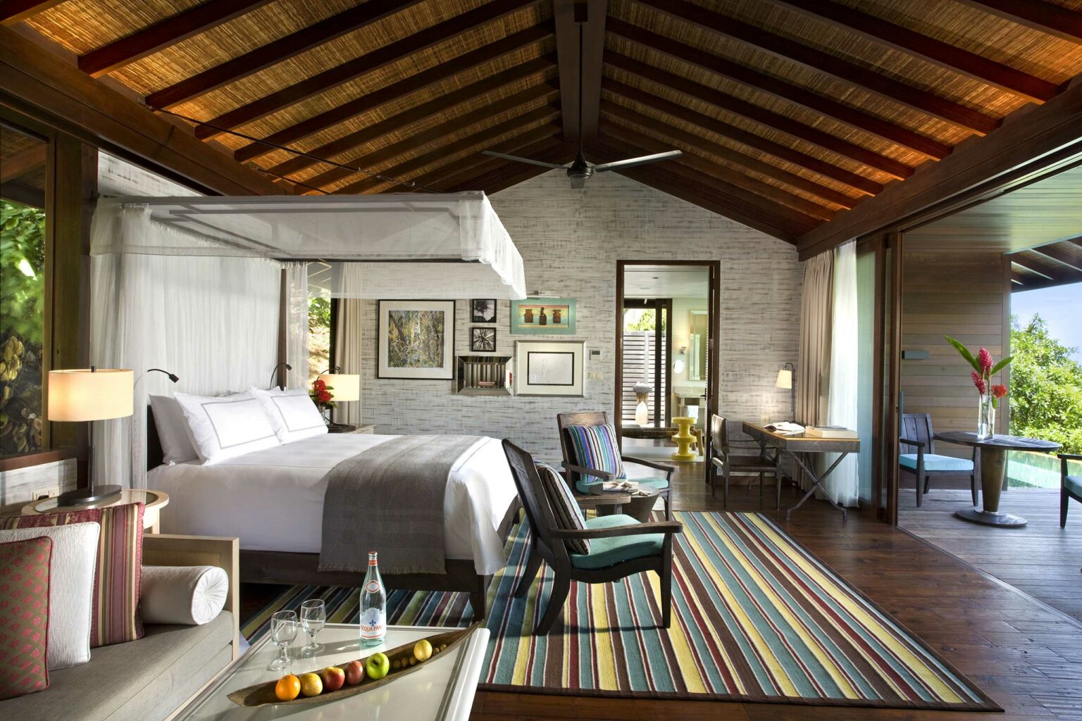 Luxurious hotel room with a canopy bed, vibrant striped rug, and open sliding doors to a terrace. Tropical greenery adds a serene touch.