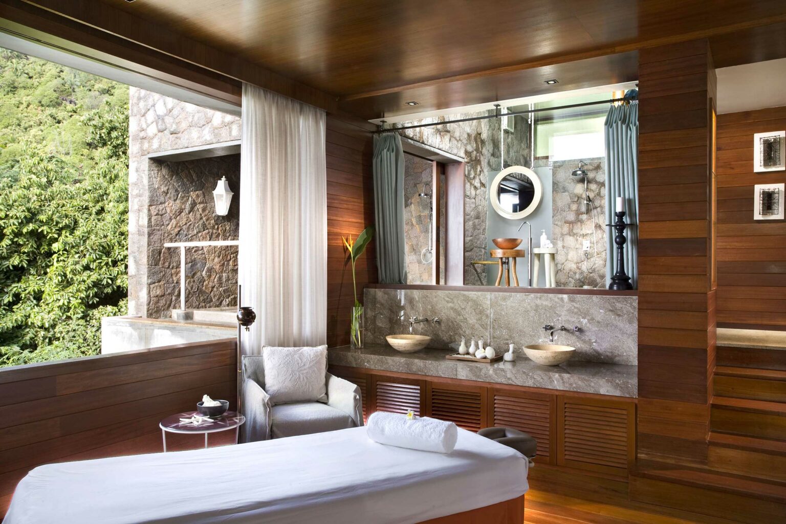 Hotel spa room with a massage table, cozy armchair, and wooden decor. A stone wall with a round mirror and candles enhances the serene, luxurious ambiance.
