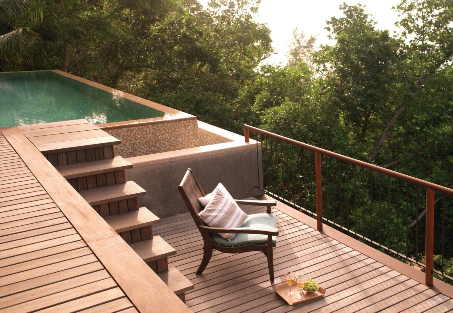 A wooden hotel deck overlooking lush greenery, featuring an infinity pool and a cushioned chair with striped pillows. Nearby, a small table holds drinks and snacks, creating a serene, inviting atmosphere.