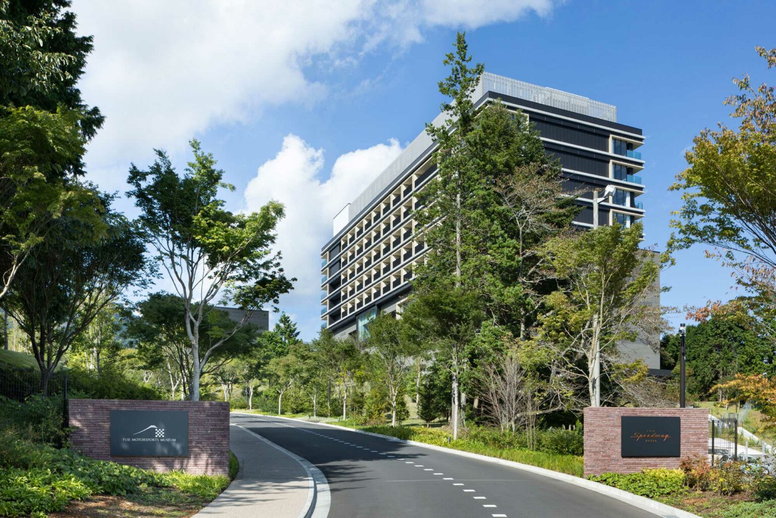 Exterior of Fuji Speedway Hotel, the Unbound Collection. It features a contemporary multi-story hotel surrounded by lush greenery and blue sky. A winding road leads to the entrance, exuding a tranquil and welcoming vibe.