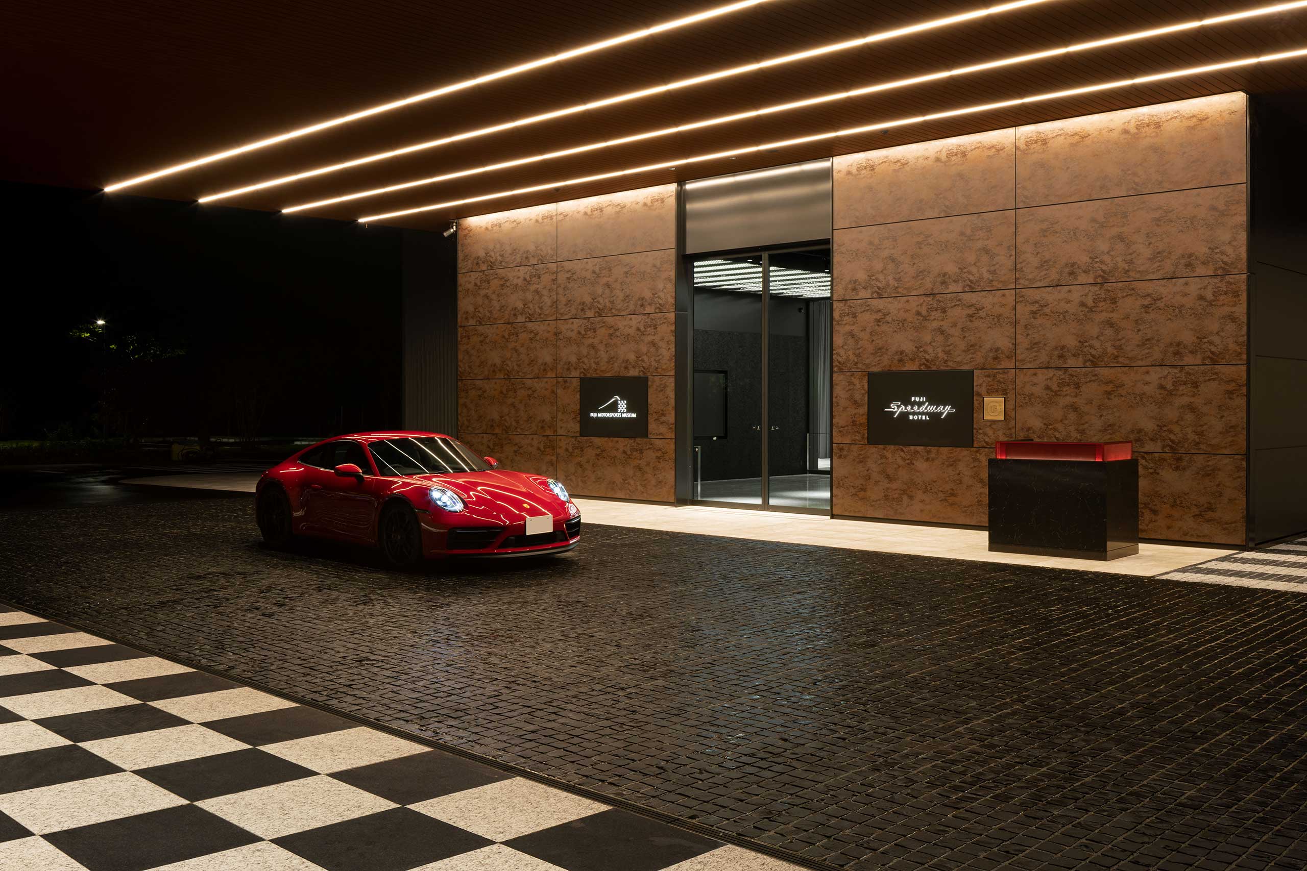 Sleek red sports car parked outside an upscale, modern hotel building at night. Warm overhead lights and textured walls create a luxurious ambiance.