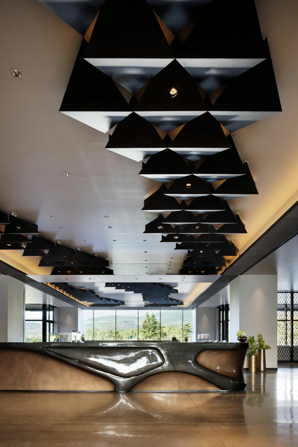 Modern hotel lobby with a sleek black reception desk. The ceiling features geometric black triangular patterns. Large windows reveal a lush, green landscape.