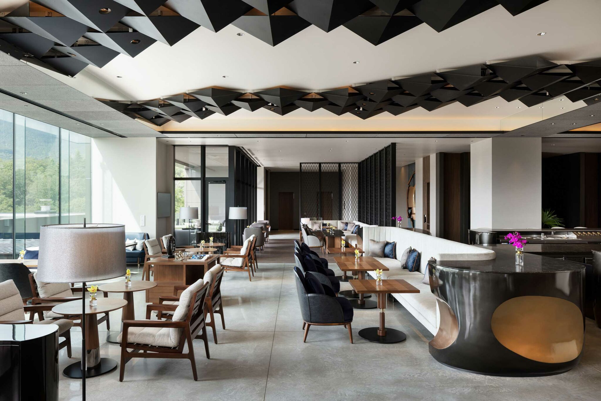 Modern hotel lobby with geometric black ceiling design. Features sleek furniture, wooden tables, and bright windows, conveying a chic and welcoming ambiance.