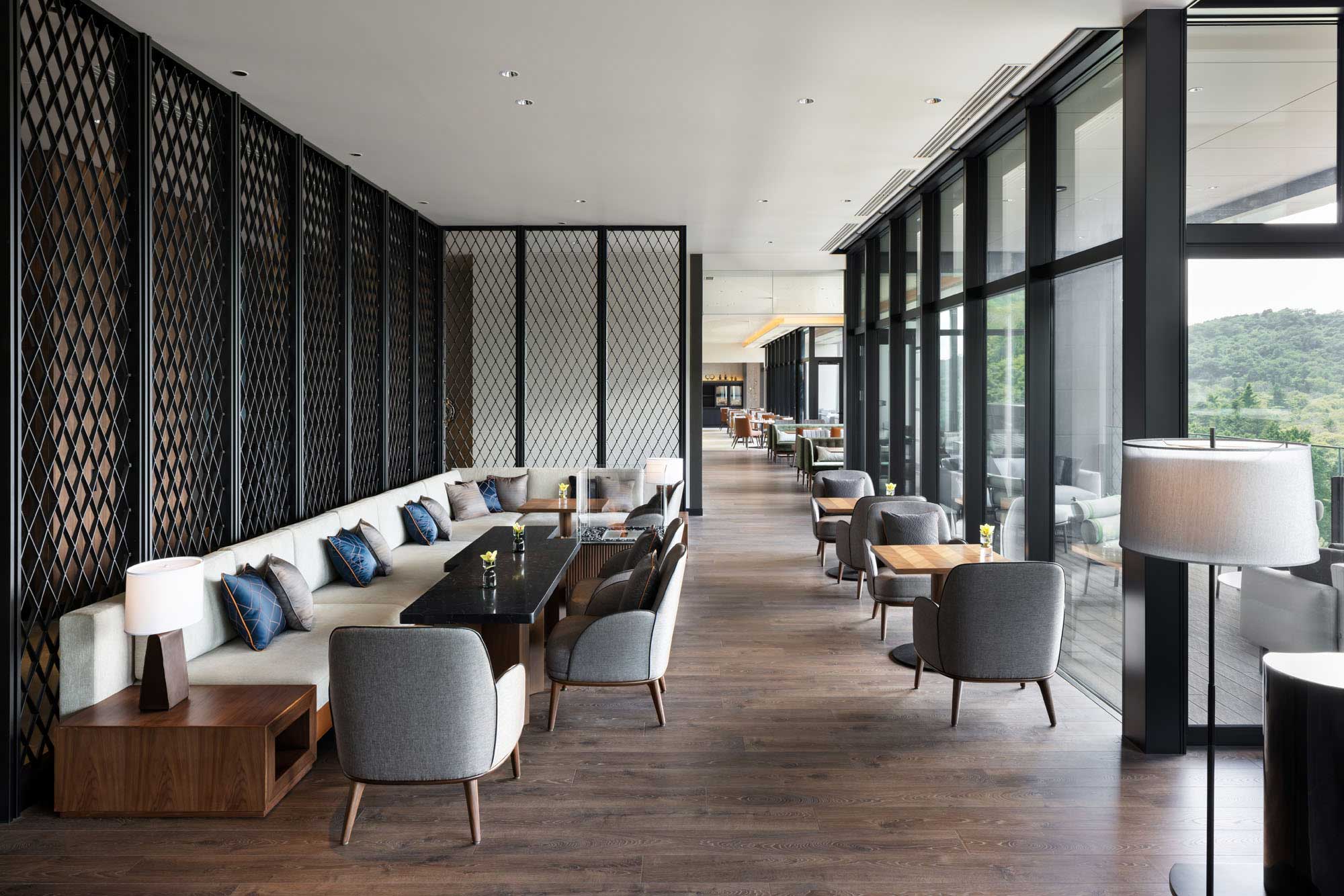 Modern hotel lounge area with a stylish interior of gray chairs, a long sofa with blue cushions, wooden tables, and black lattice screens. Large windows overlook greenery.