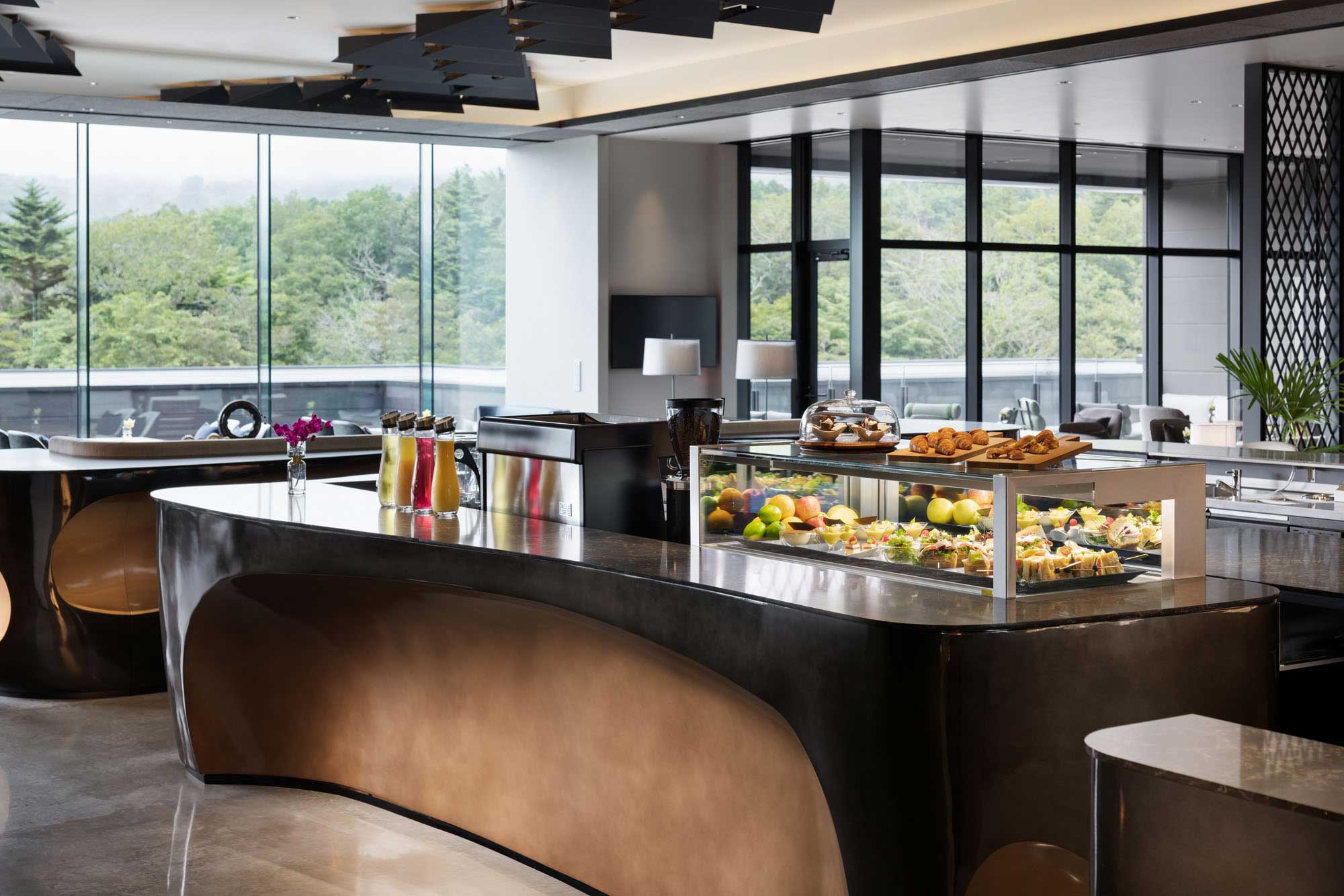 Sleek modern hotel dining area with curved counters and a display case filled with pastries and fruit. Large windows offer views of lush greenery outside.