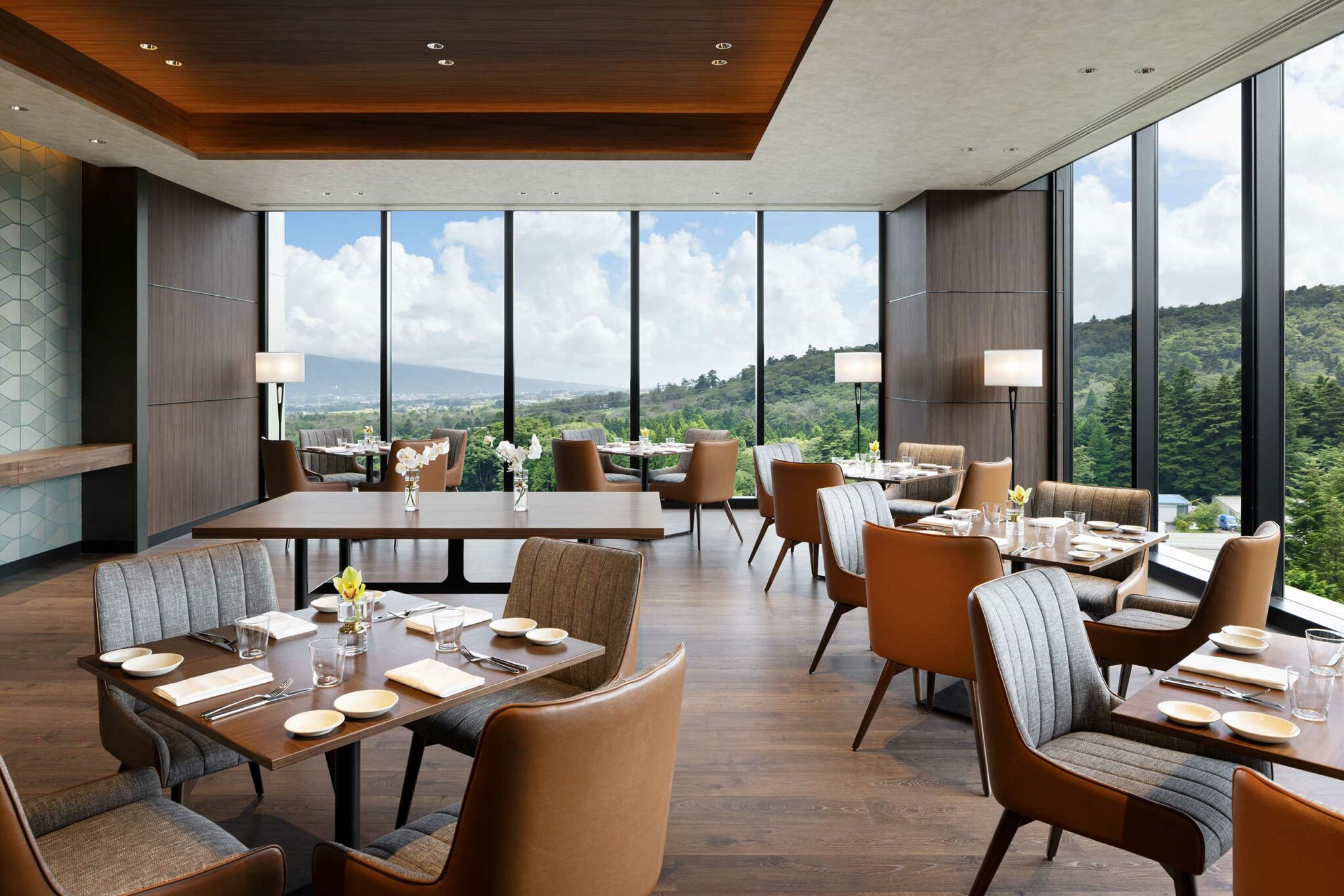 Modern hotel restaurant with large windows offers a scenic view of hills and cloudy skies. Elegant tables are set with neutral-toned chairs and decor.