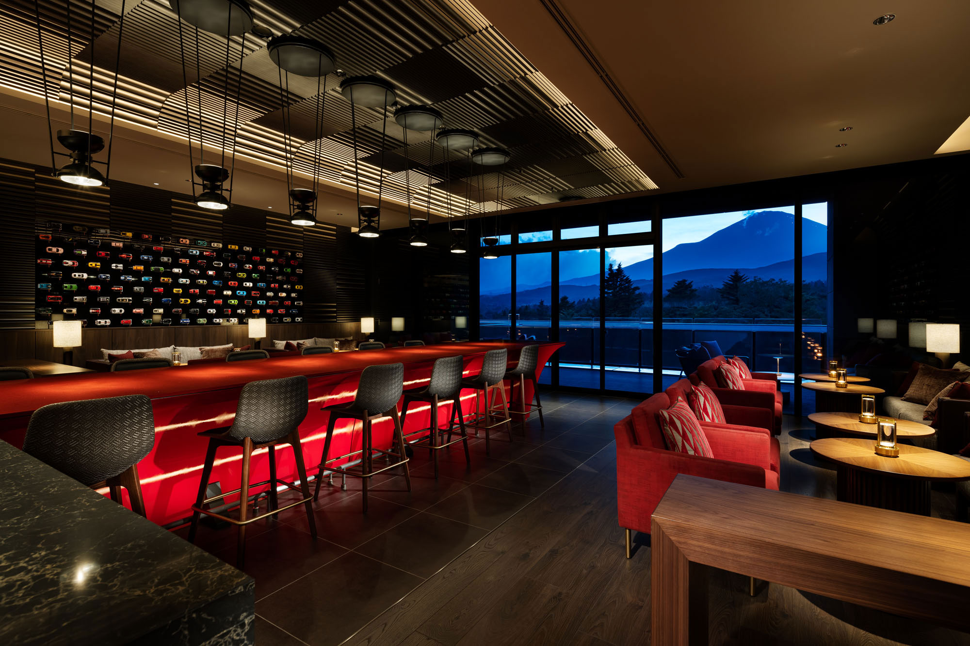 A stylish, dimly lit hotel bar with a sleek red counter and gray high chairs. Large windows reveal a serene mountain view at dusk, creating a cozy ambiance.