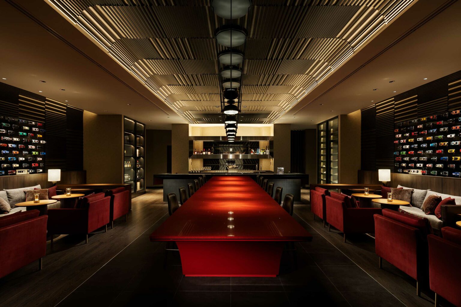 A modern, elegant hotel restaurant interior with a long red table in the center, surrounded by red chairs. Soft lighting and wine bottles create a cozy ambiance.