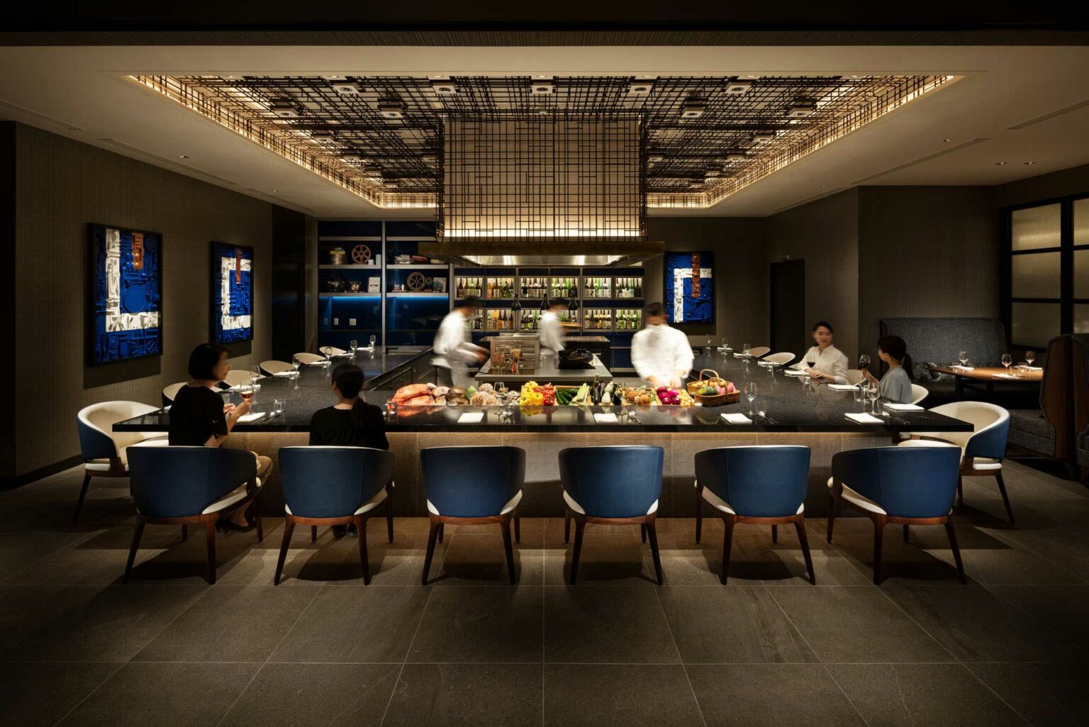 Modern hotel restaurant scene with a sleek bar counter adorned with colorful fruits. Chefs in white coats prepare dishes, while patrons dine under warm ambient lighting.