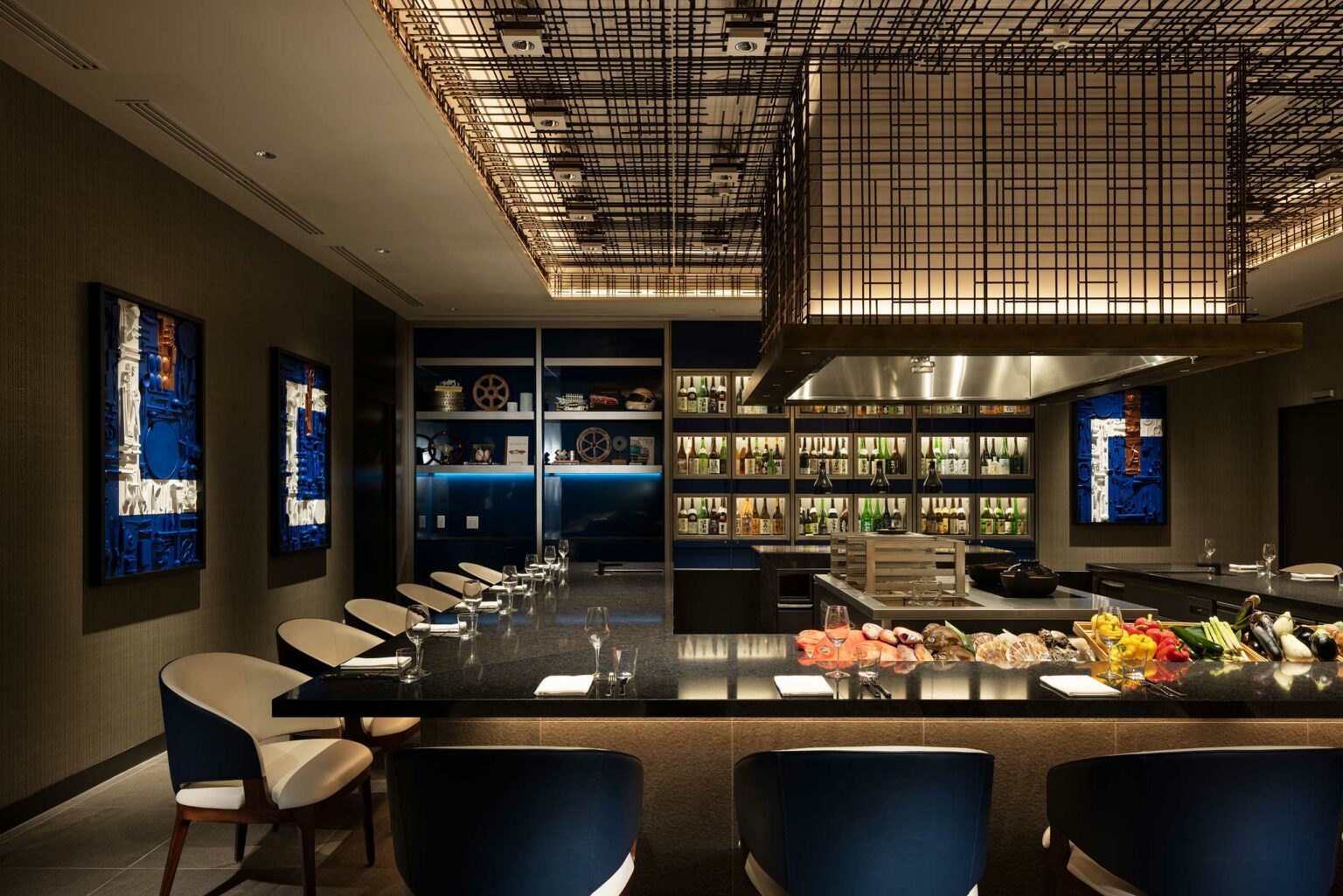 Sophisticated hotel dining area with a sleek black counter, elegant cushioned chairs, decorative wall art, and a well-lit ceiling. Bottles line shelves in the background.