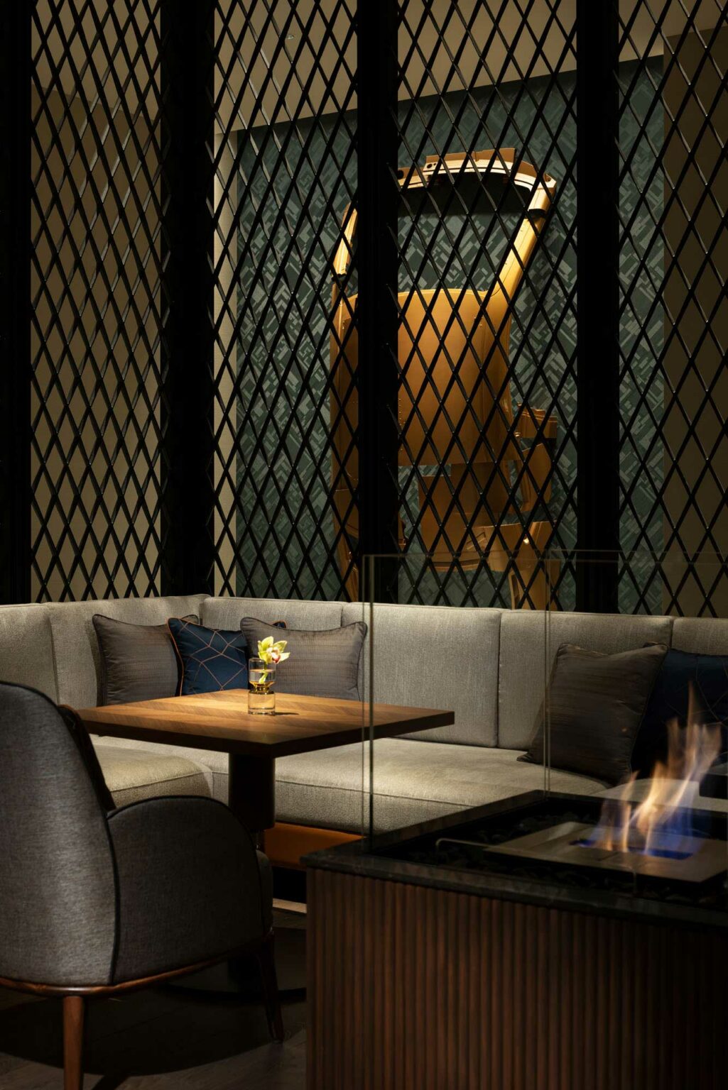 Cozy hotel indoor seating area with a grey sectional sofa, wooden table, and small plant. A warm fire burns in a sleek glass fireplace, creating a relaxing ambiance.