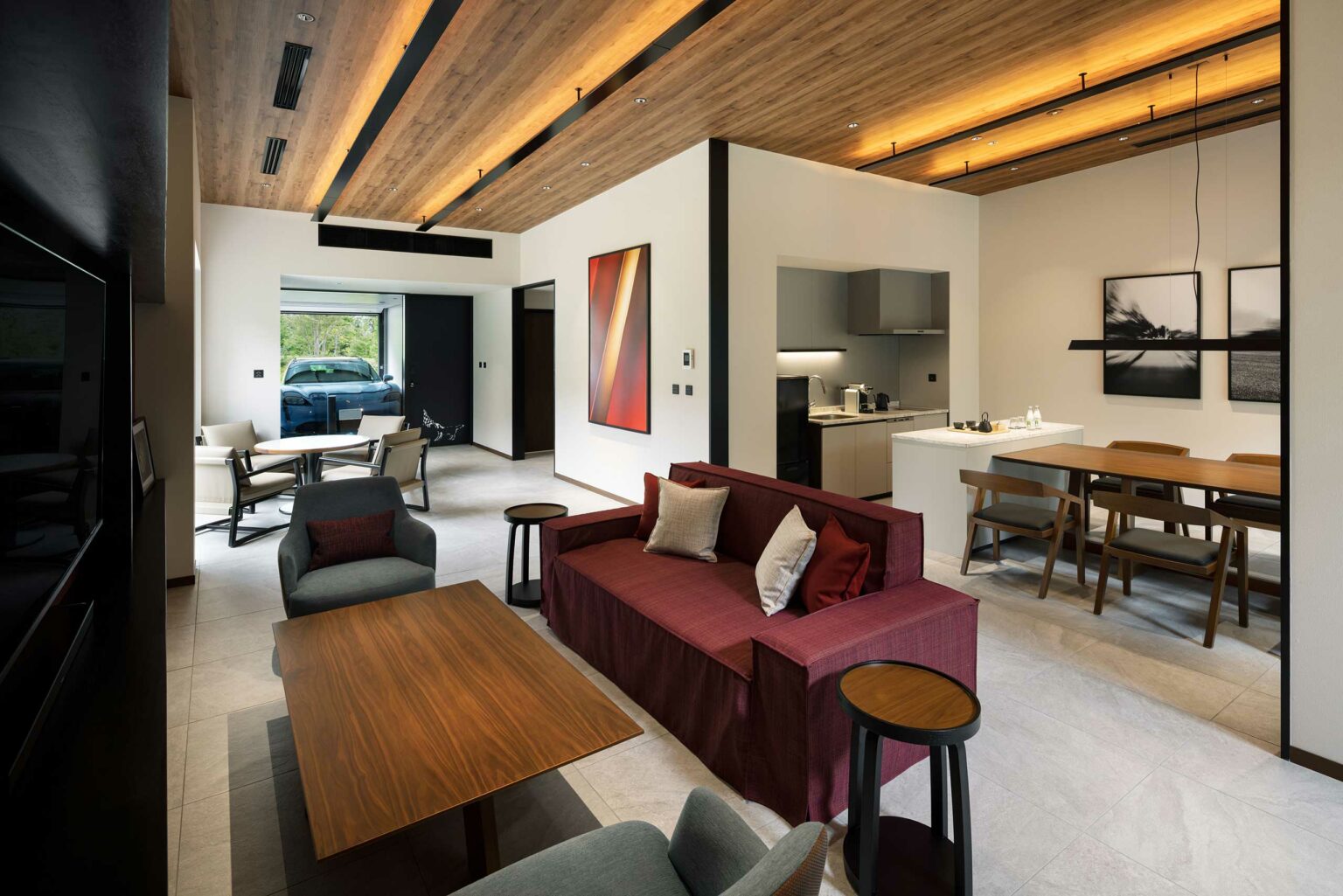 Modern guest room living area featuring a burgundy sofa with cushions, a wooden ceiling, abstract artwork, and a kitchen with a cozy dining space. Warm, inviting ambiance.
