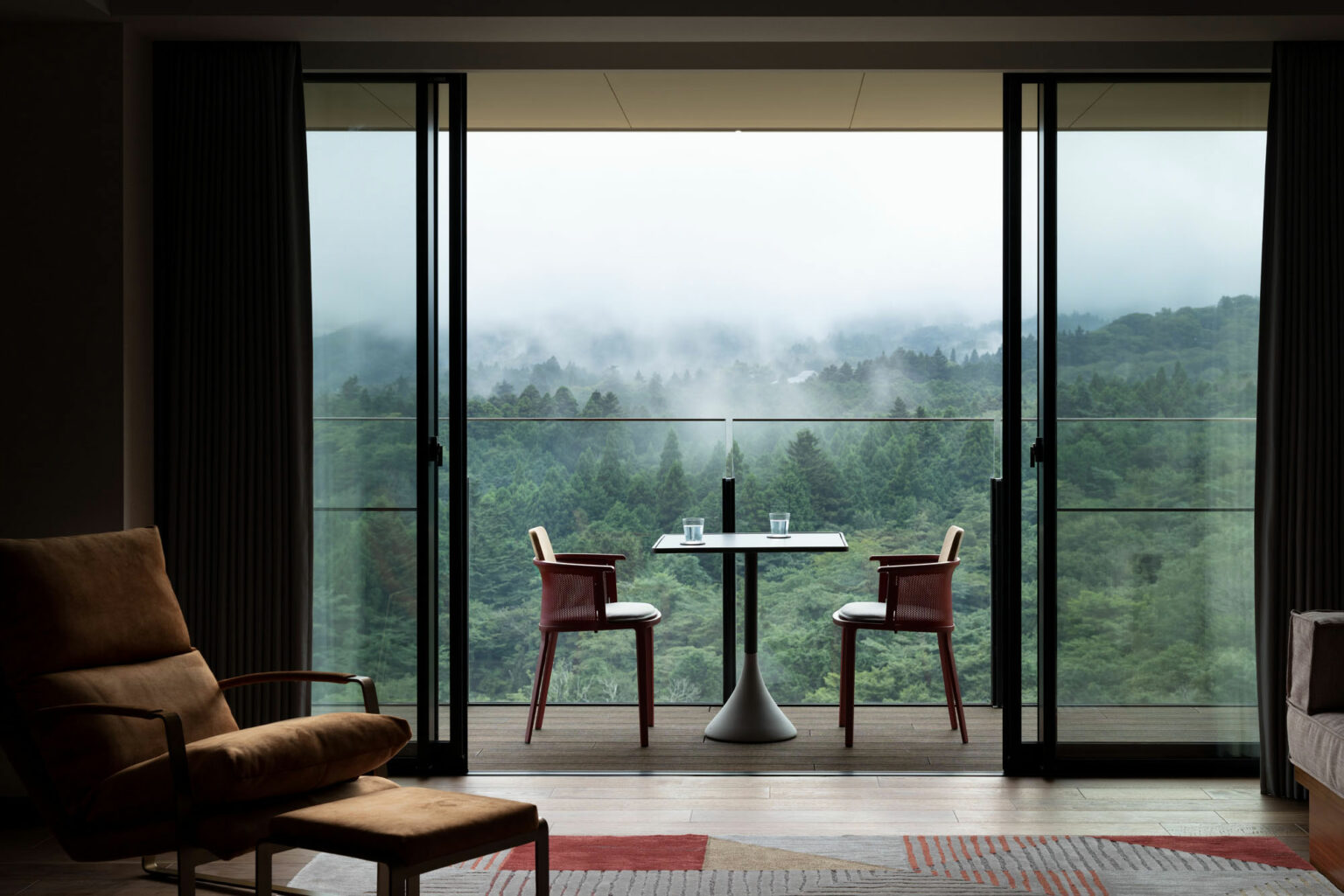Cozy hotel bedroom with an open balcony offering a misty forest view. A small table with two chairs and glasses, conveying tranquility and relaxation.