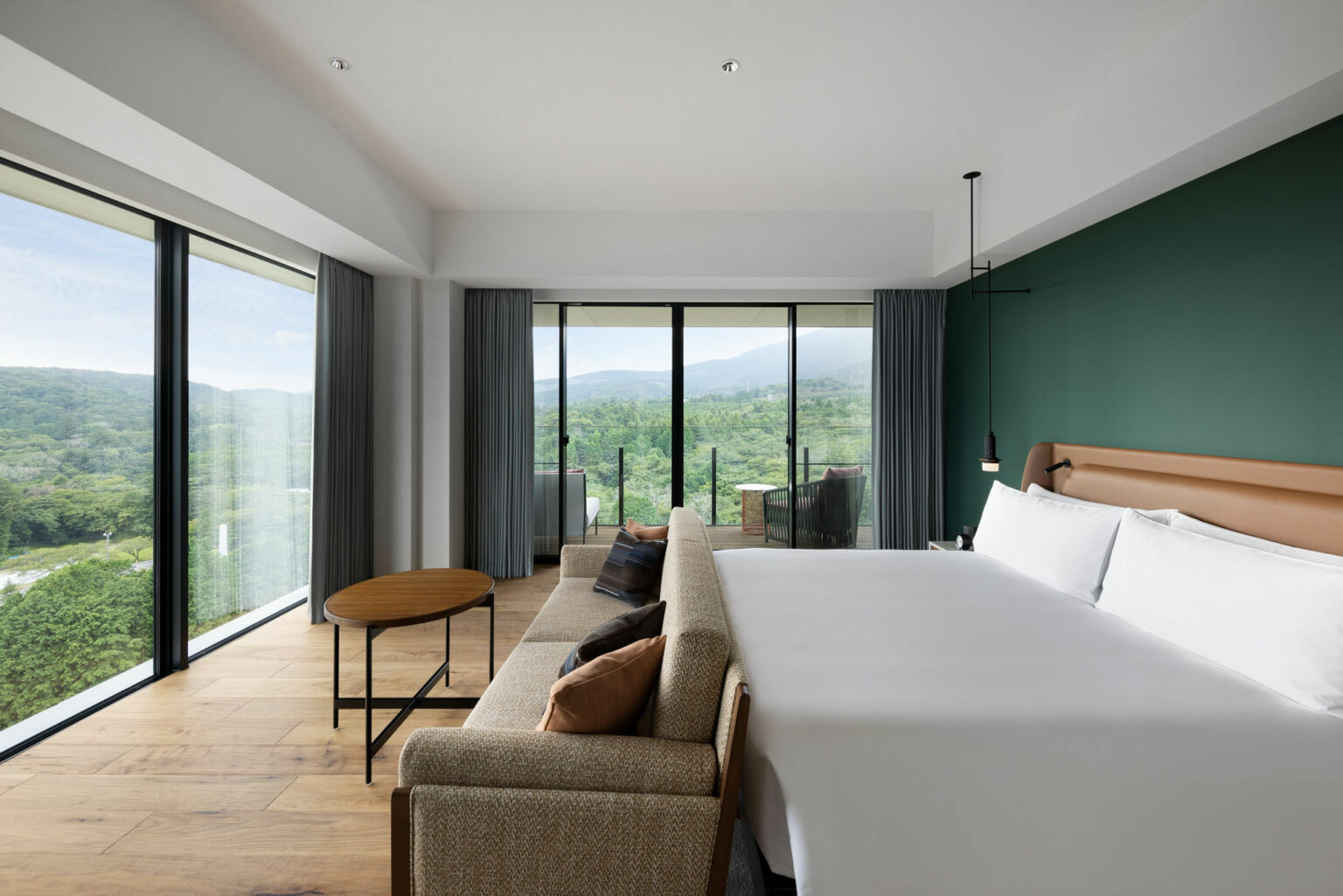 Modern hotel room with a large bed, green accent wall, and floor-to-ceiling windows offering a lush mountain view. Calm and inviting atmosphere.