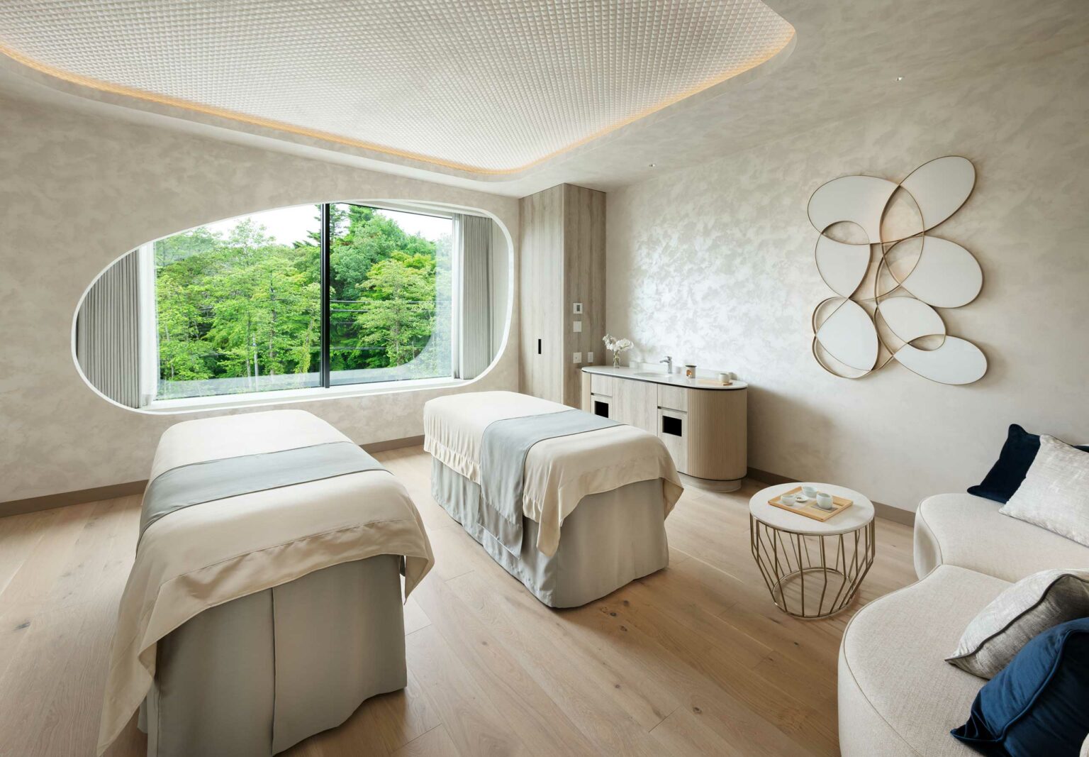 A serene hotel spa treatment room with two massage beds, soft beige decor, and a large window showcasing green trees. Modern wall art and calming ambiance prevail.