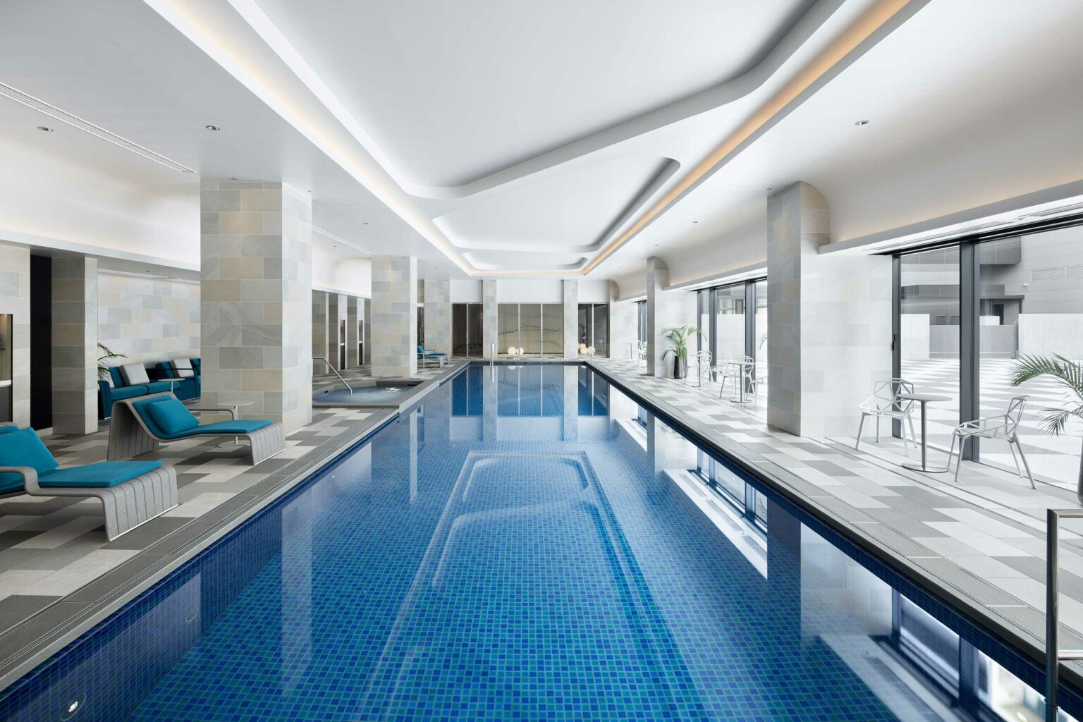 Spacious indoor hotel swimming pool with blue tiles and chic lounge chairs. Natural light streams in through large windows, creating a serene, luxurious ambiance.
