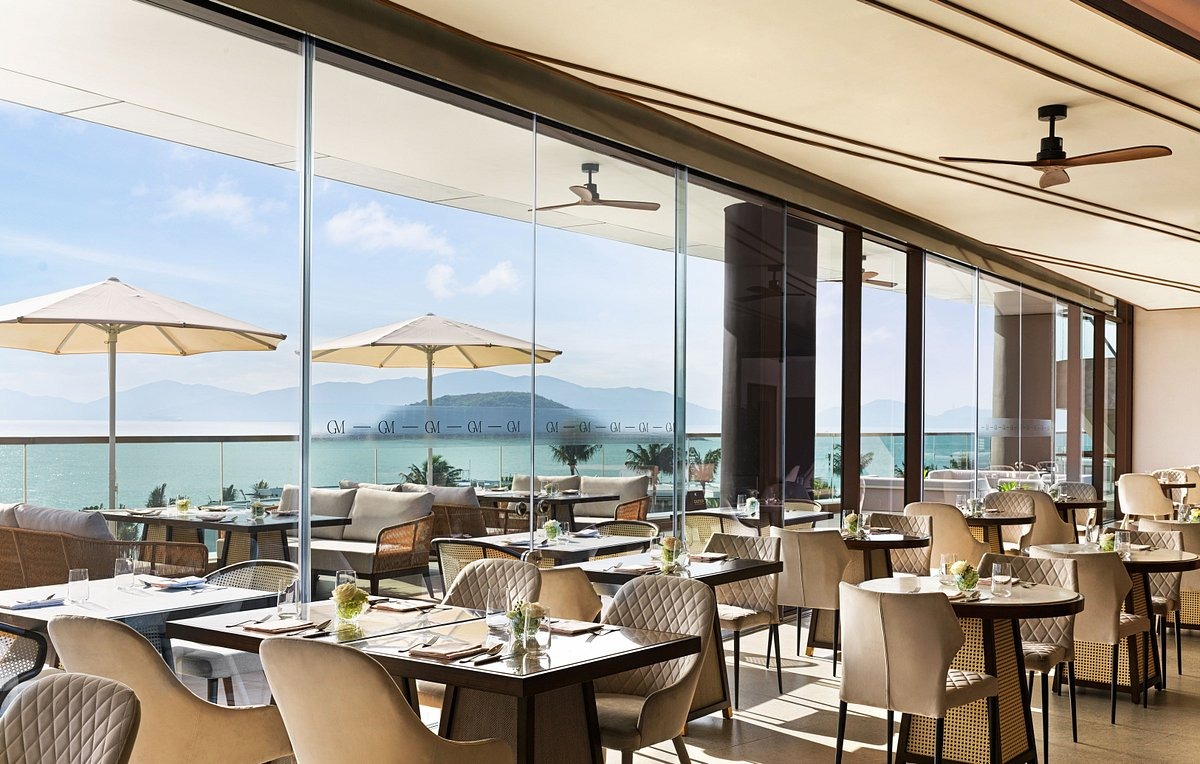 Luxurious, airy hotel cafe with ocean view. Features wicker chairs, green bar stools, potted plants, modern decor, and a server arranging items.