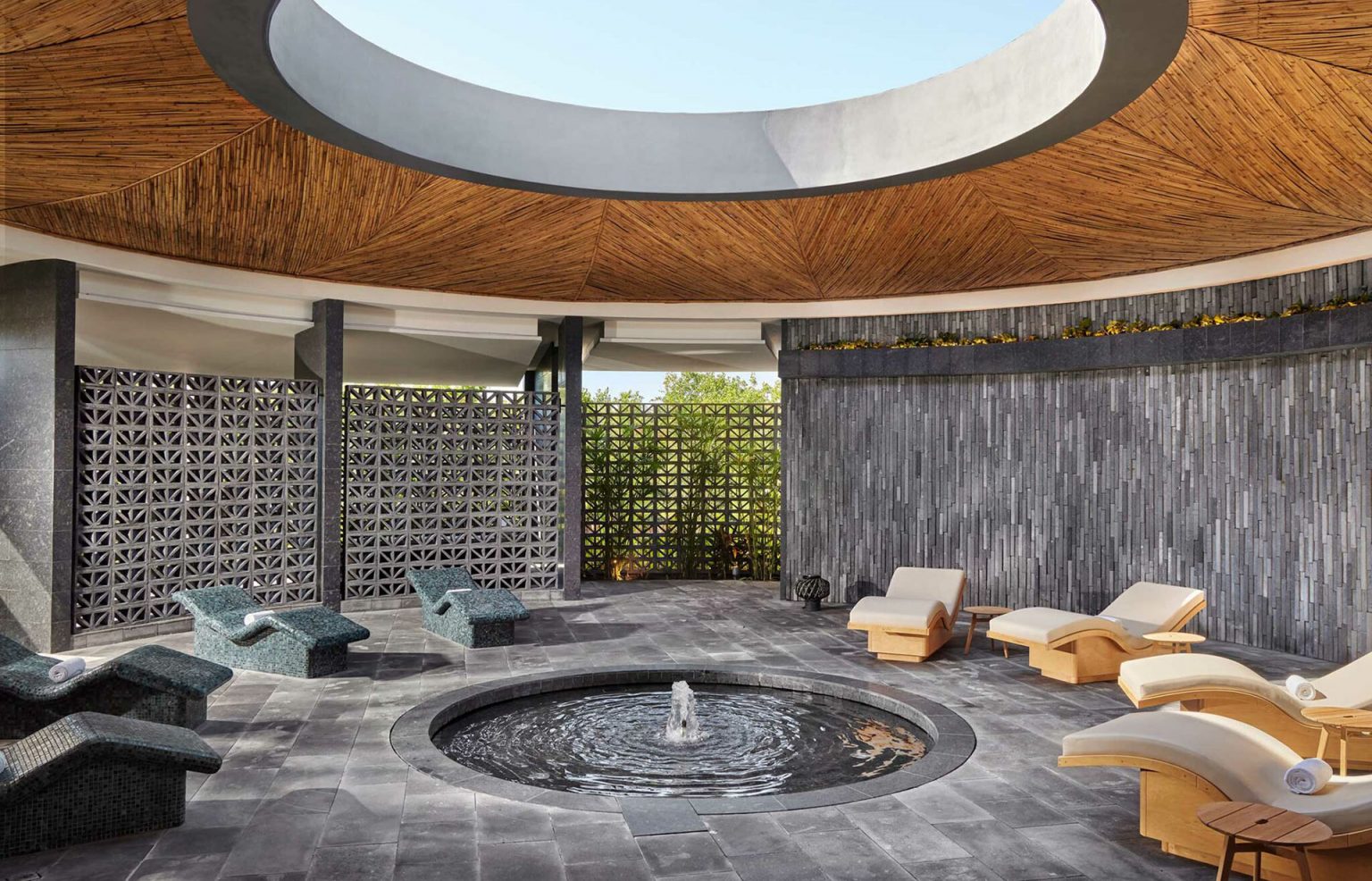 Spacious resort spa interior with a circular skylight, bamboo ceiling, modern loungers, and a central water feature. The ambiance is serene and luxurious.
