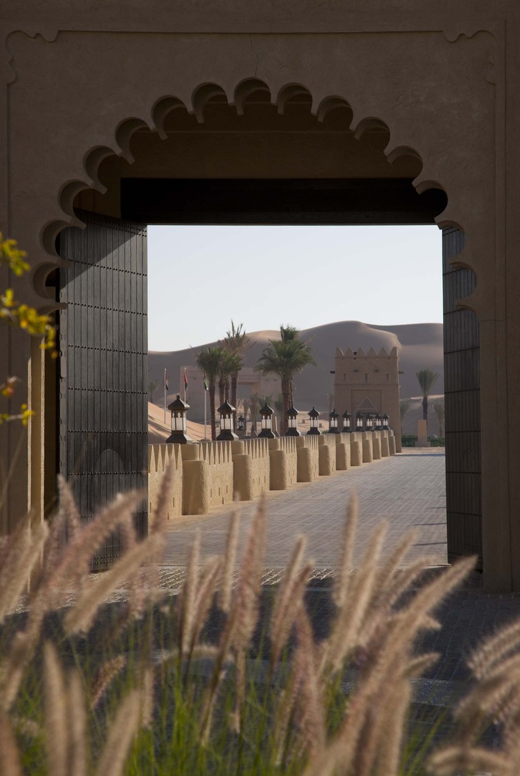 A historic sand-colored fort with tall, cylindrical towers reflects in a tranquil pool. Palm trees and a spouting fountain enhance the serene scene.