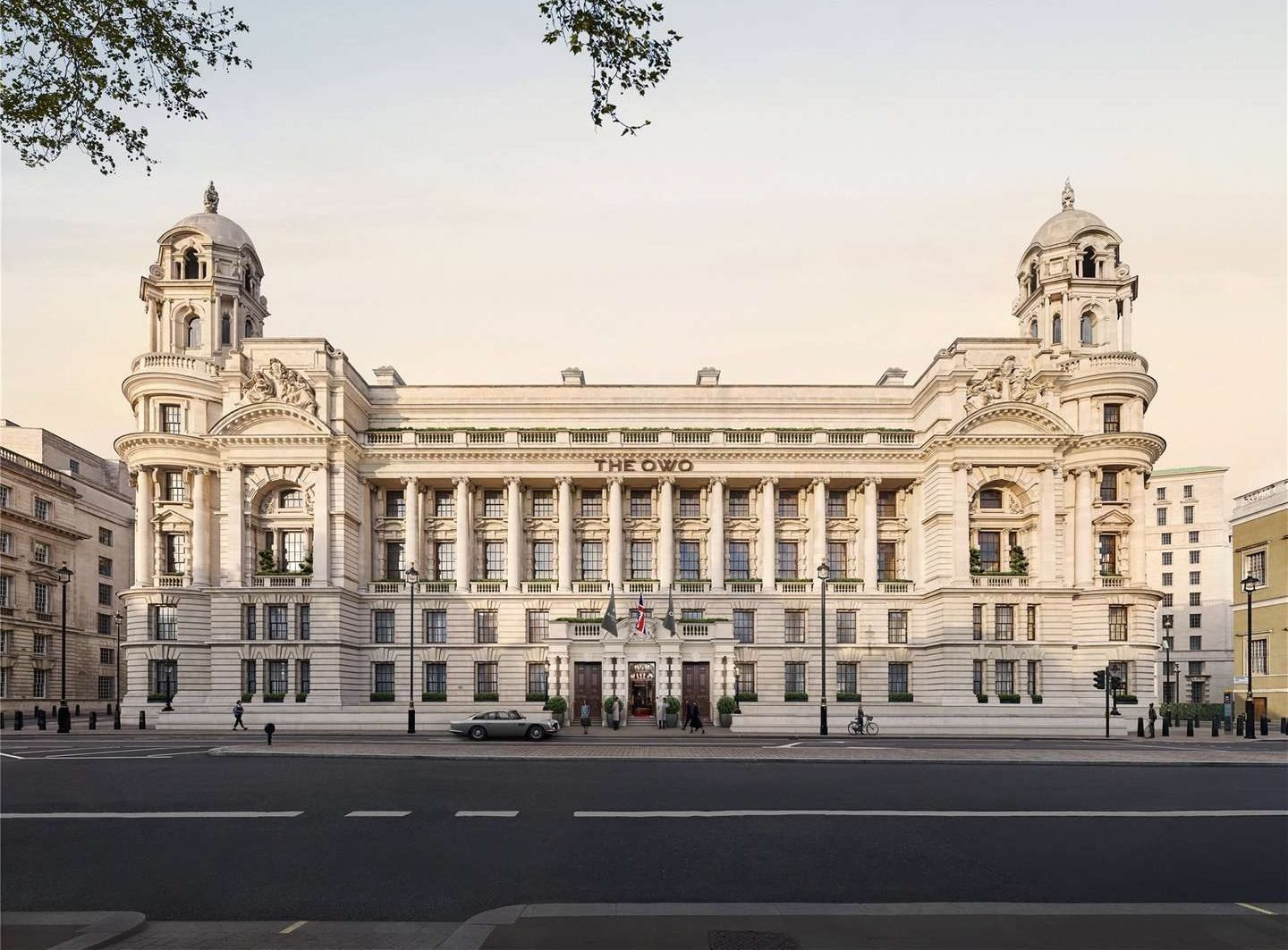 The OWO Residences by Raffles exteriors. Majestic neoclassical building with grand columns, ornate facades, and two towers; the setting is serene with a pale sky and sparse traffic.