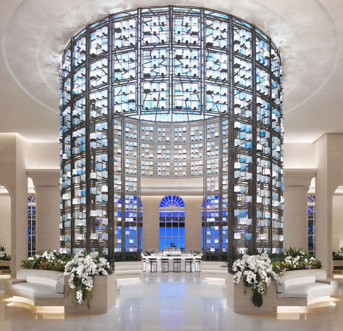 Luxurious hotel lobby featuring a towering cylindrical glass wine display with soft blue lighting, lush white floral arrangements, and elegant seating.