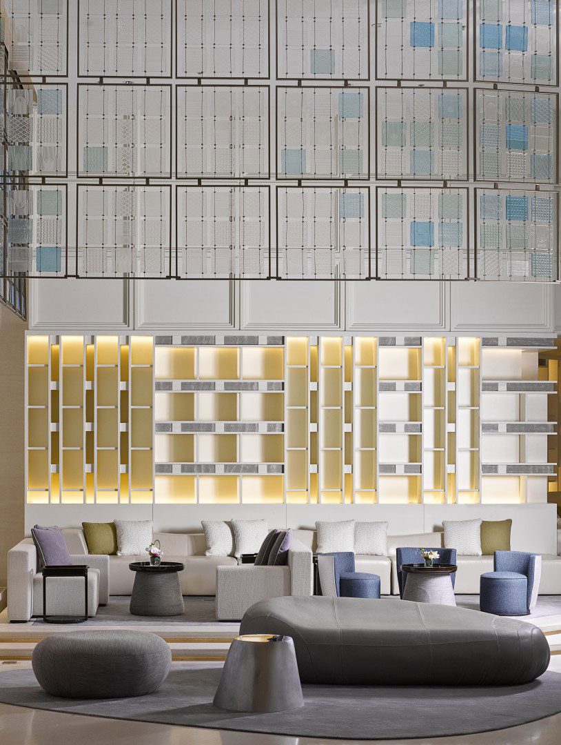 Contemporary hotel lounge with geometric wall panels, backlit shelves, cushioned seating, and minimalist tables. Soft, neutral tones create a tranquil atmosphere.