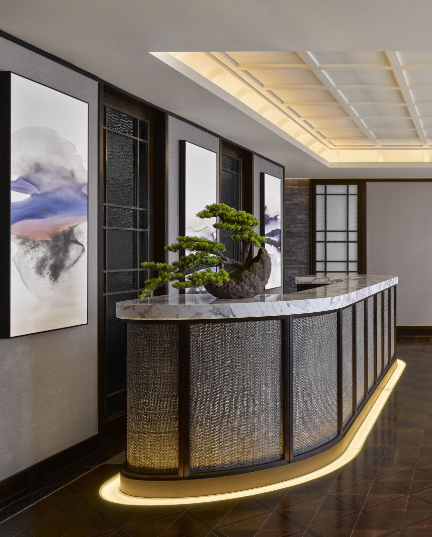 A modern hotel reception desk with a sleek marble top and illuminated base stands under a coffered ceiling. A bonsai tree decorates the desk, flanked by abstract art on the walls. The ambiance is elegant and tranquil.