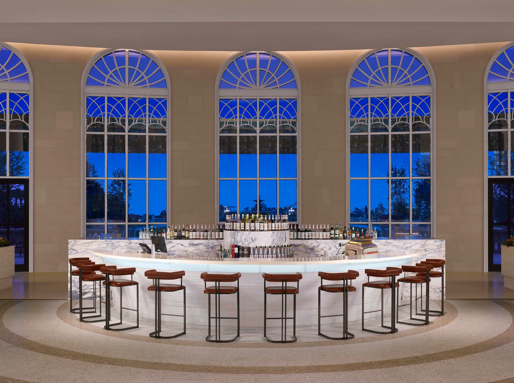 Elegant curved marble bar with high stools, set against large arched windows showing a twilight view within a hotel. Bottles line the bar, conveying a sophisticated ambiance.