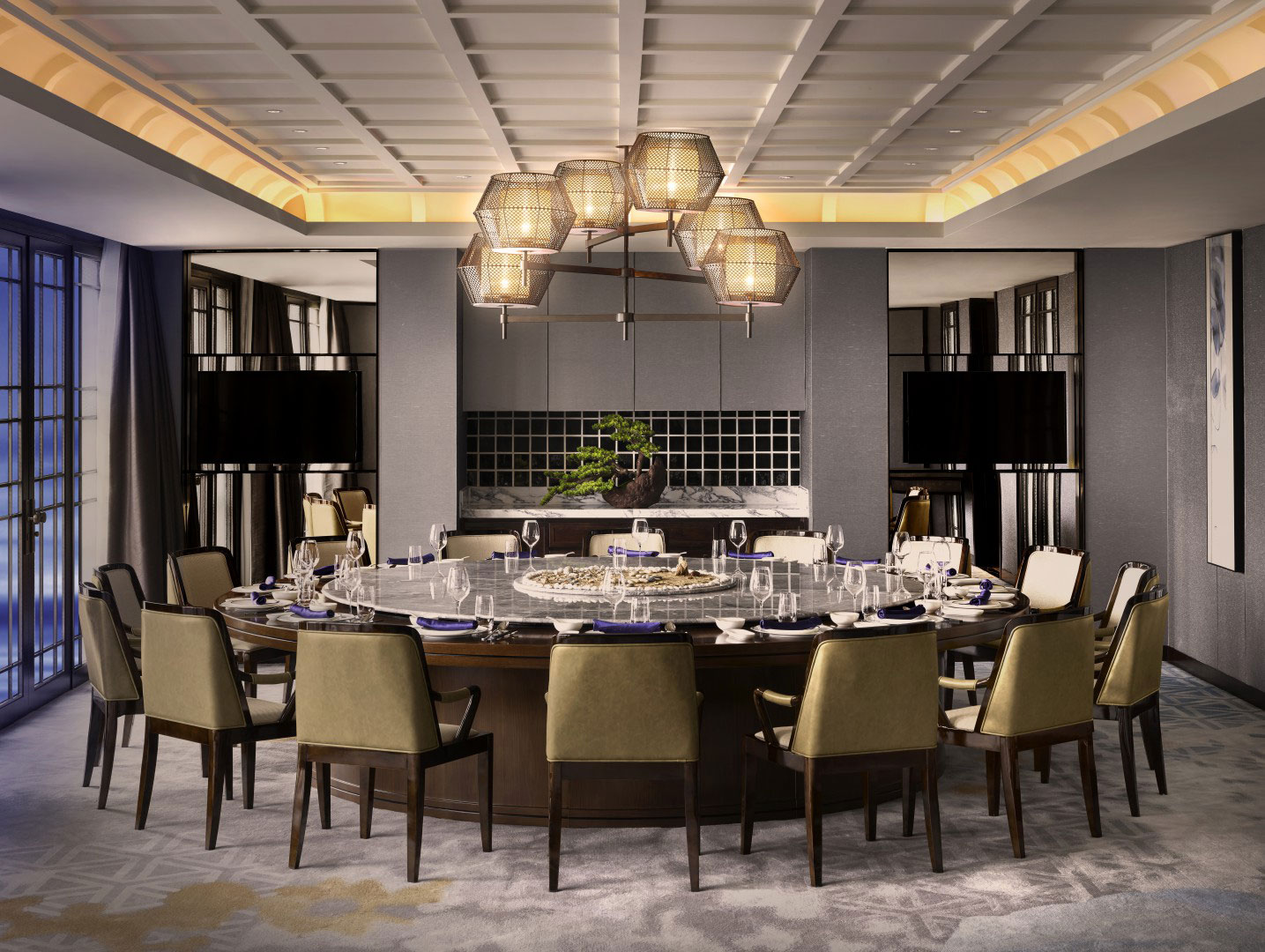 Elegant hotel dining room with a large round table set for a meal, surrounded by cushioned chairs. Modern chandelier above; minimalist decor with a serene bonsai on a side table.