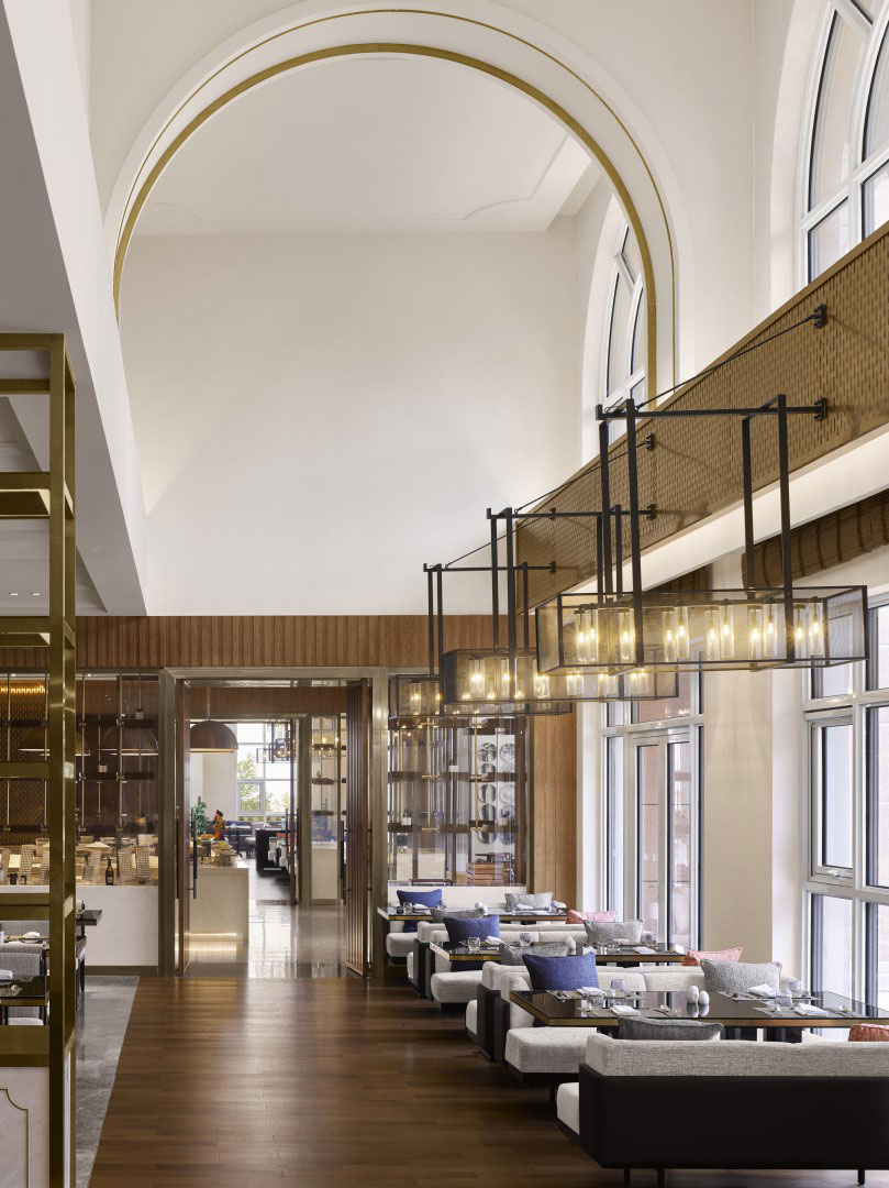 Elegant hotel restaurant interior with high arched ceilings, large windows, modern lighting, and neatly arranged tables with blue and pink cushions. Luxurious ambiance.