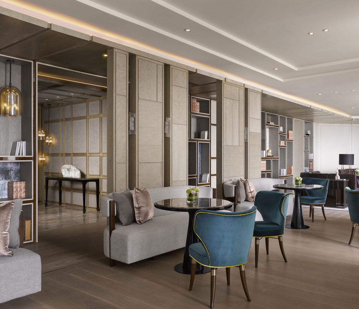 Modern hotel lounge with elegant decor, featuring grey sofas, blue velvet chairs, and round tables. Soft lighting and bookshelves create a cozy ambiance.