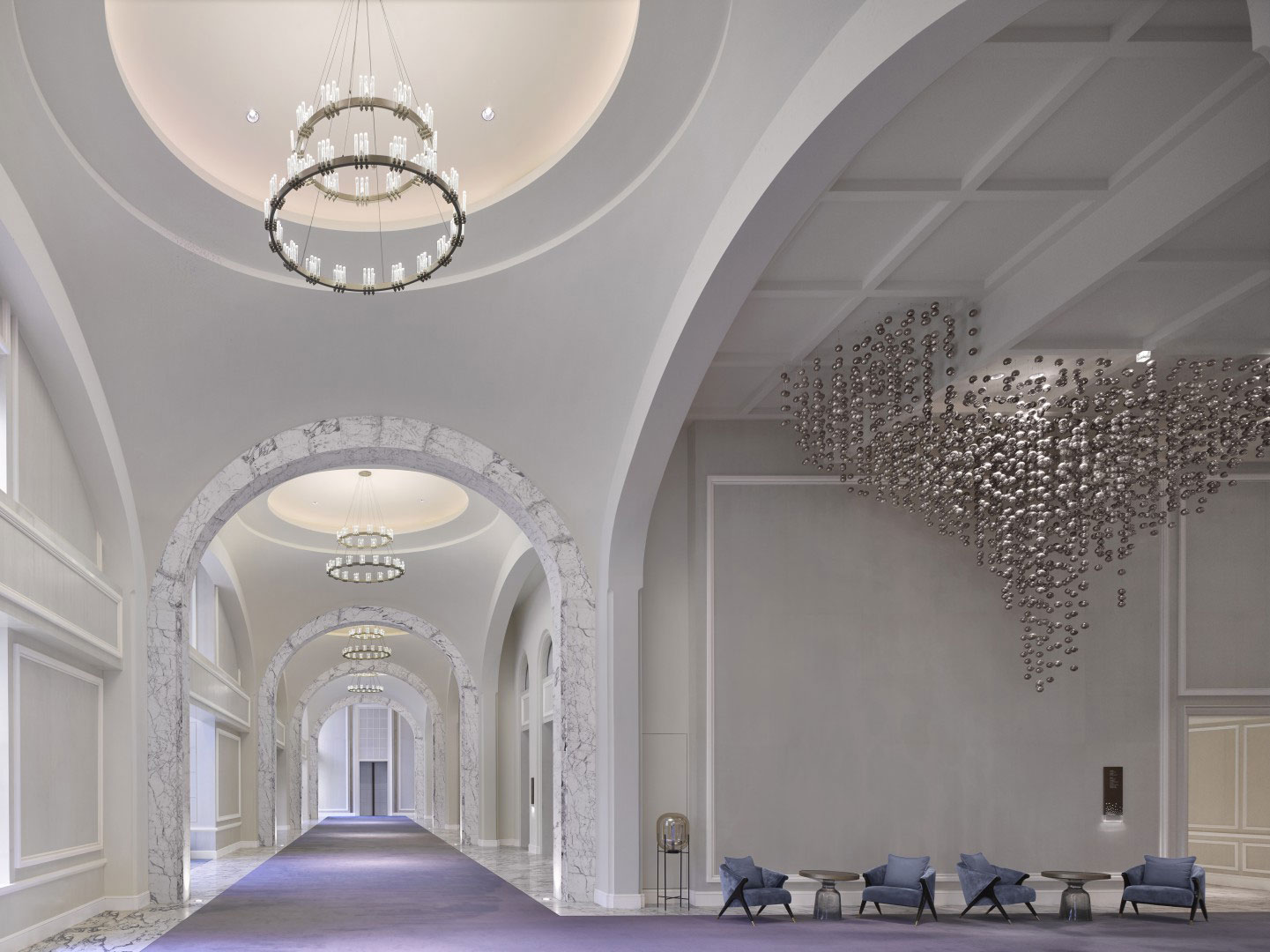 Elegant hotel corridor with marble arches, large chandeliers, and a shimmering wall sculpture. Blue carpet and chairs create a luxurious, serene ambiance.