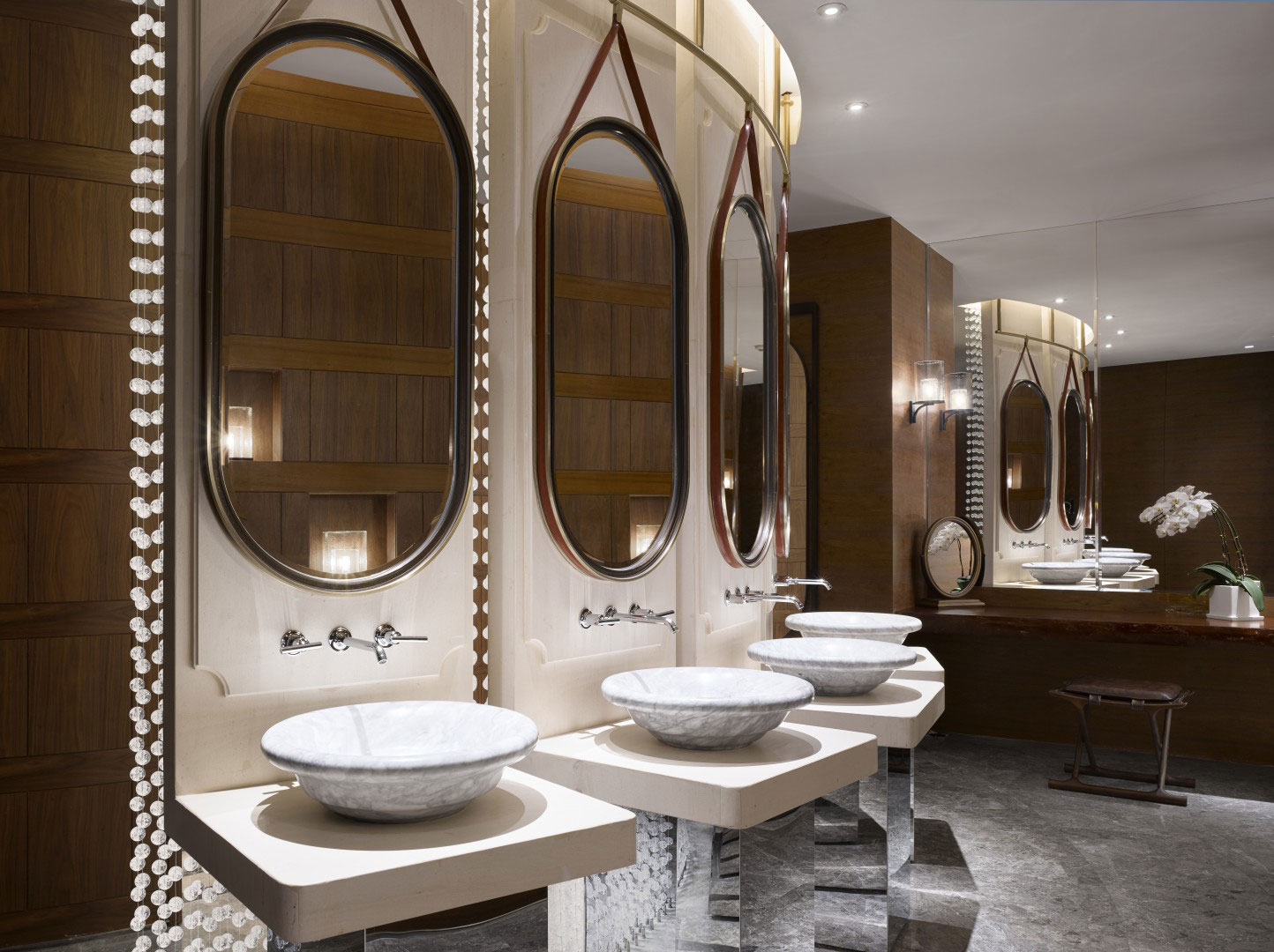 Elegant hotel spa restroom with four white marble sinks and oval mirrors on a beige wall. Warm lighting and wood accents create a sophisticated, serene atmosphere.
