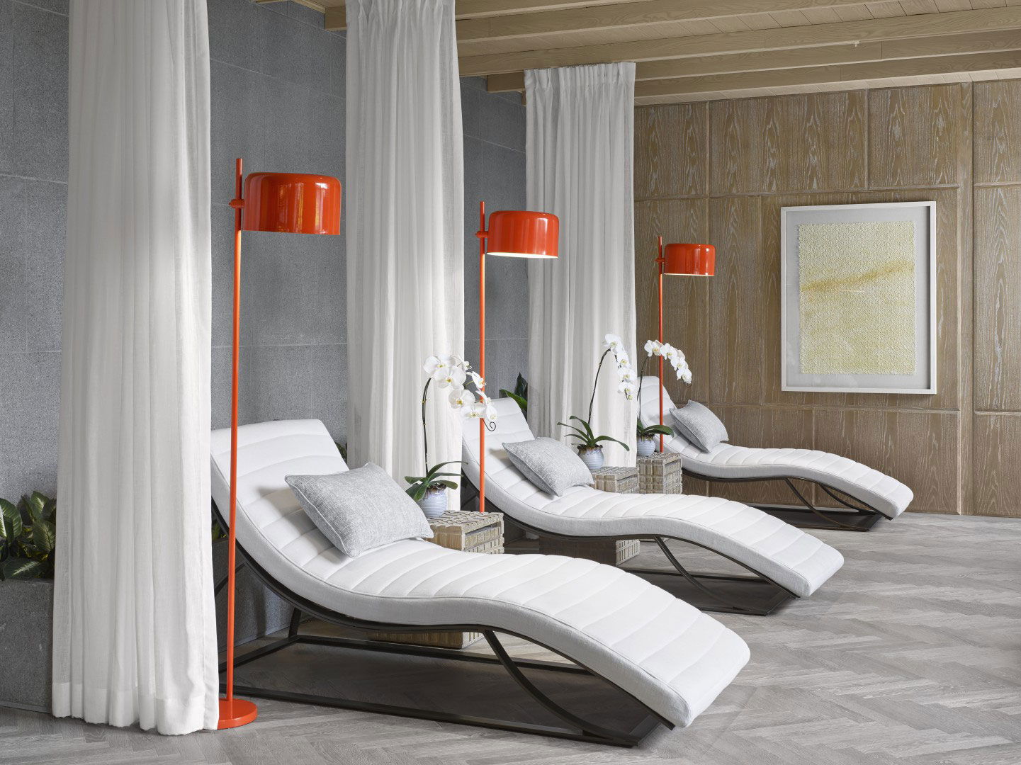 Modern hotel spa lounge with three white curved chaise lounges, gray pillows, and vibrant orange floor lamps. Orchids and elegant wood paneling add a serene touch.