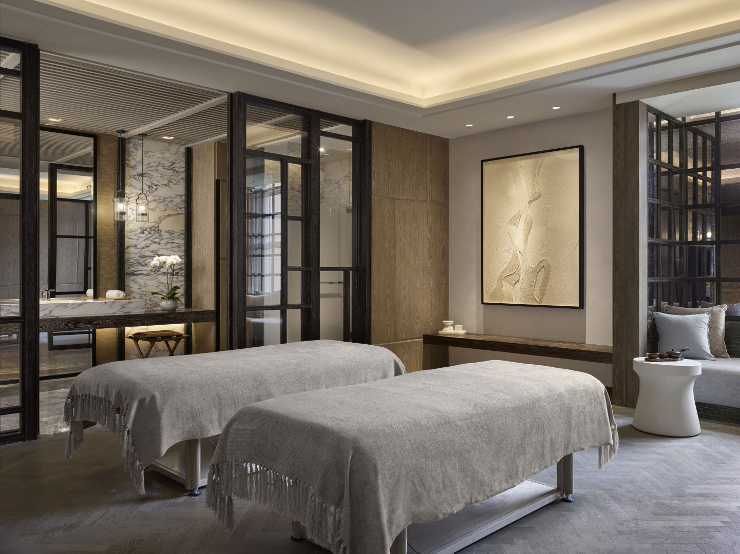 Calm hotel spa treatment room with two massage beds covered in light gray linens, soft lighting, abstract wall art, and a serene, sophisticated ambiance.