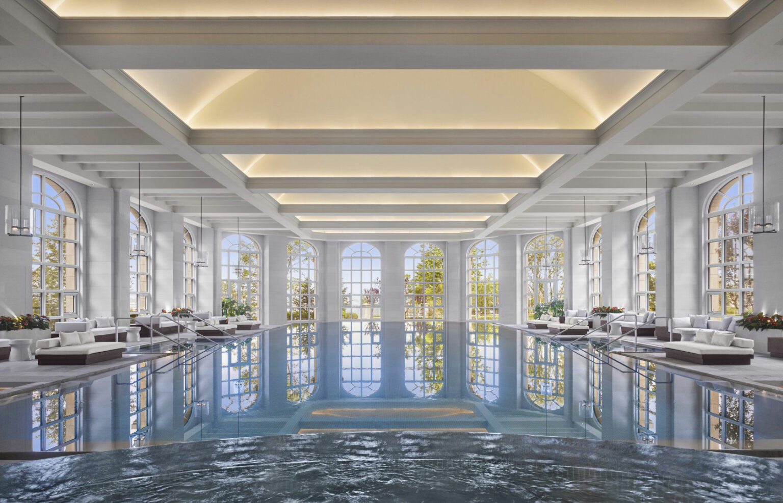 Luxurious hotel indoor pool with high arched windows, reflecting soft natural light. Elegant lounge chairs line the sides, conveying a serene and opulent atmosphere.