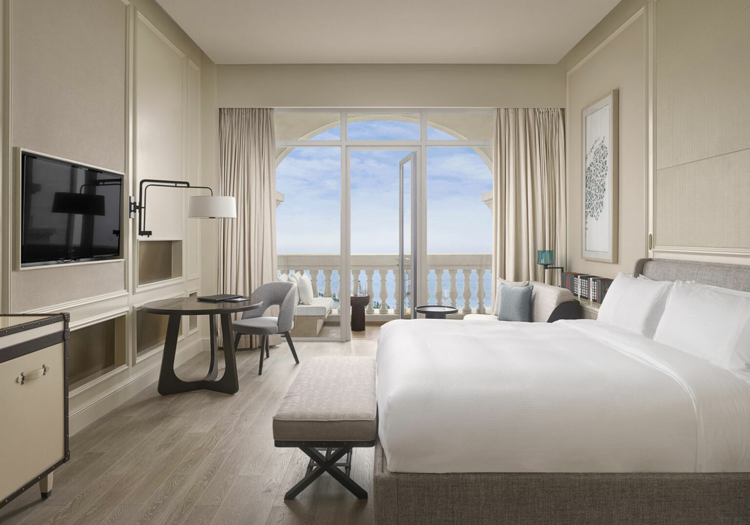 Hotel room with elegant decor, featuring a large bed, TV, and seating area. A balcony with ocean view is seen through glass doors. Calm and luxurious atmosphere.