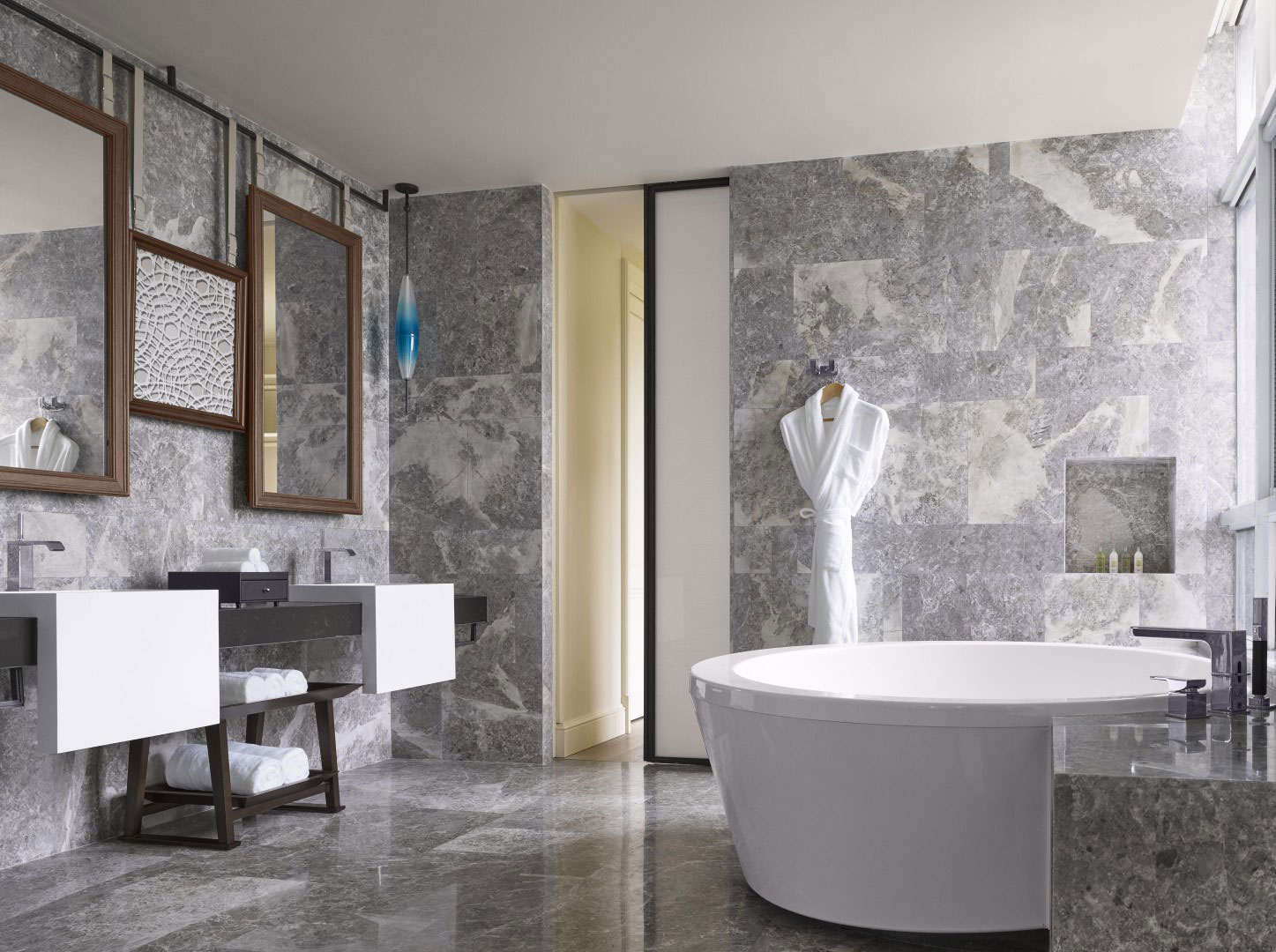 Luxurious suite bathroom with marble walls and floors, featuring a round bathtub, modern sinks, large mirrors, a hanging robe, and a serene ambiance.
