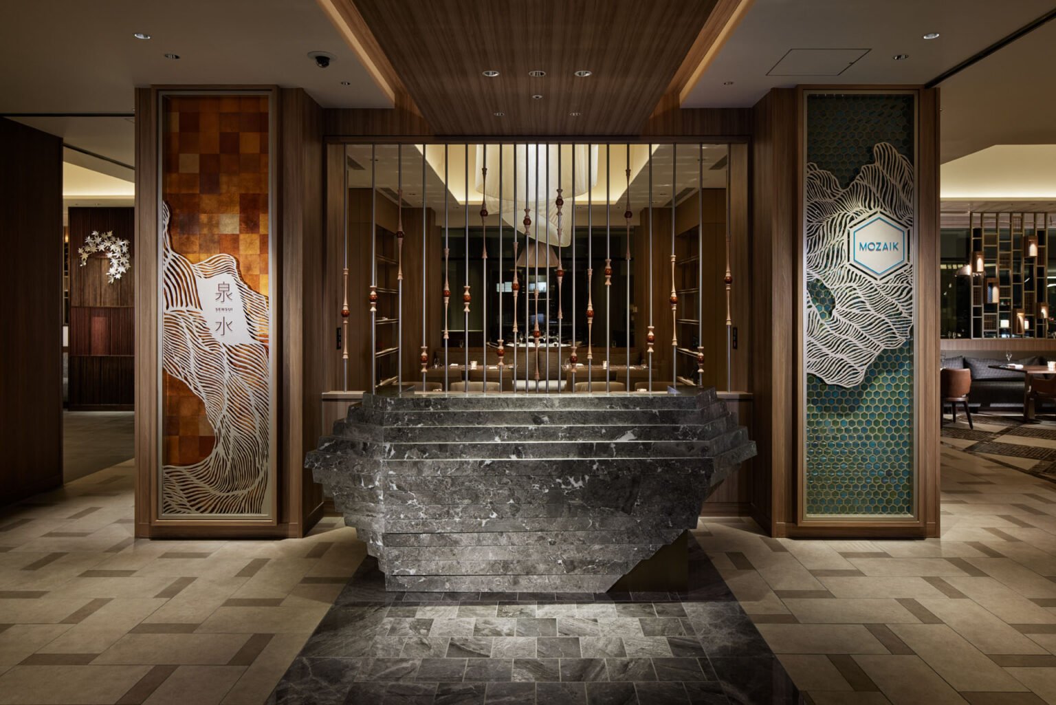 Luxurious hotel lobby with a modern, artistic design. A central stone sculpture stands before vertical rods; warm lighting and intricate wall patterns create an elegant ambiance.