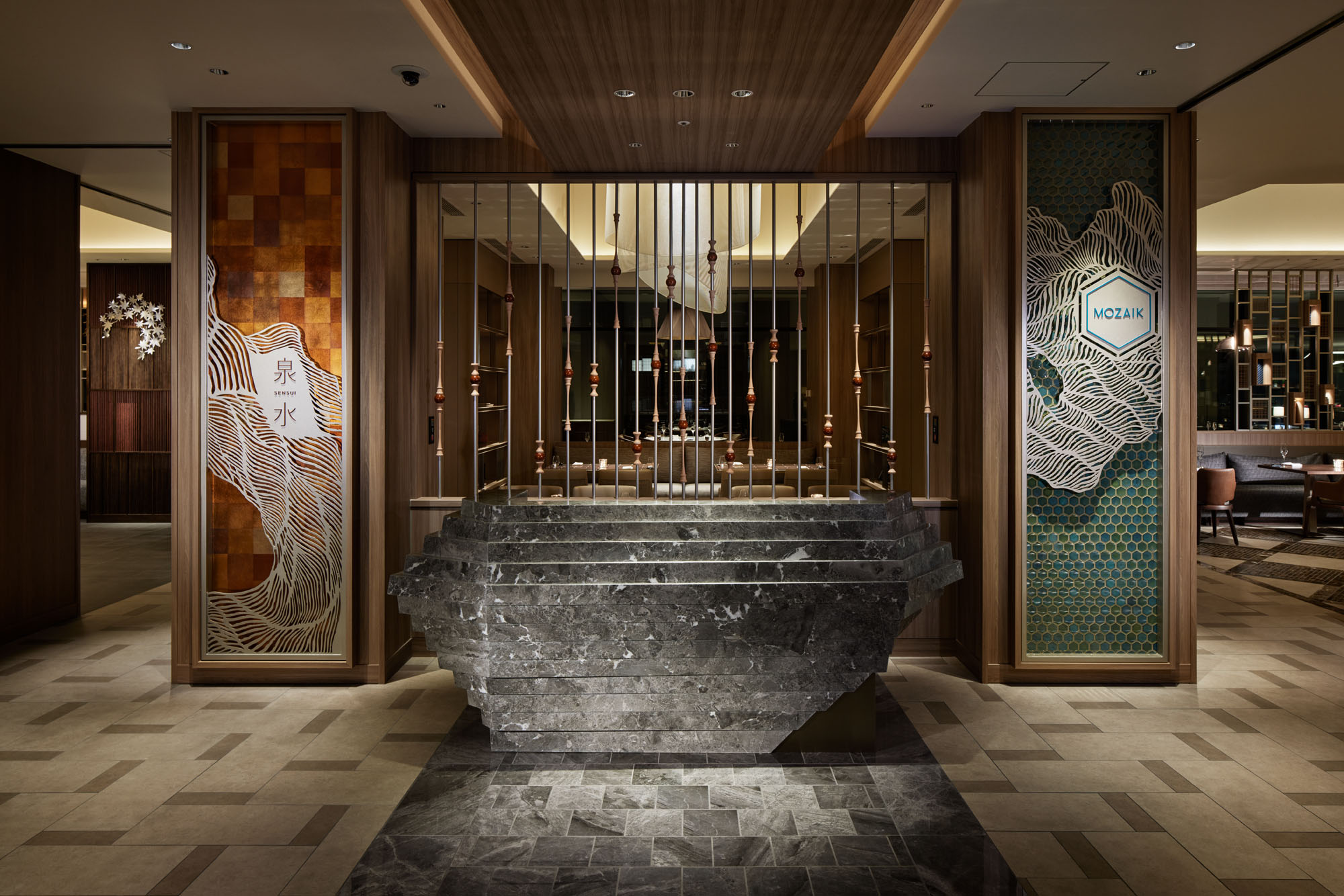 Luxurious hotel lobby with a modern, artistic design. A central stone sculpture stands before vertical rods; warm lighting and intricate wall patterns create an elegant ambiance.