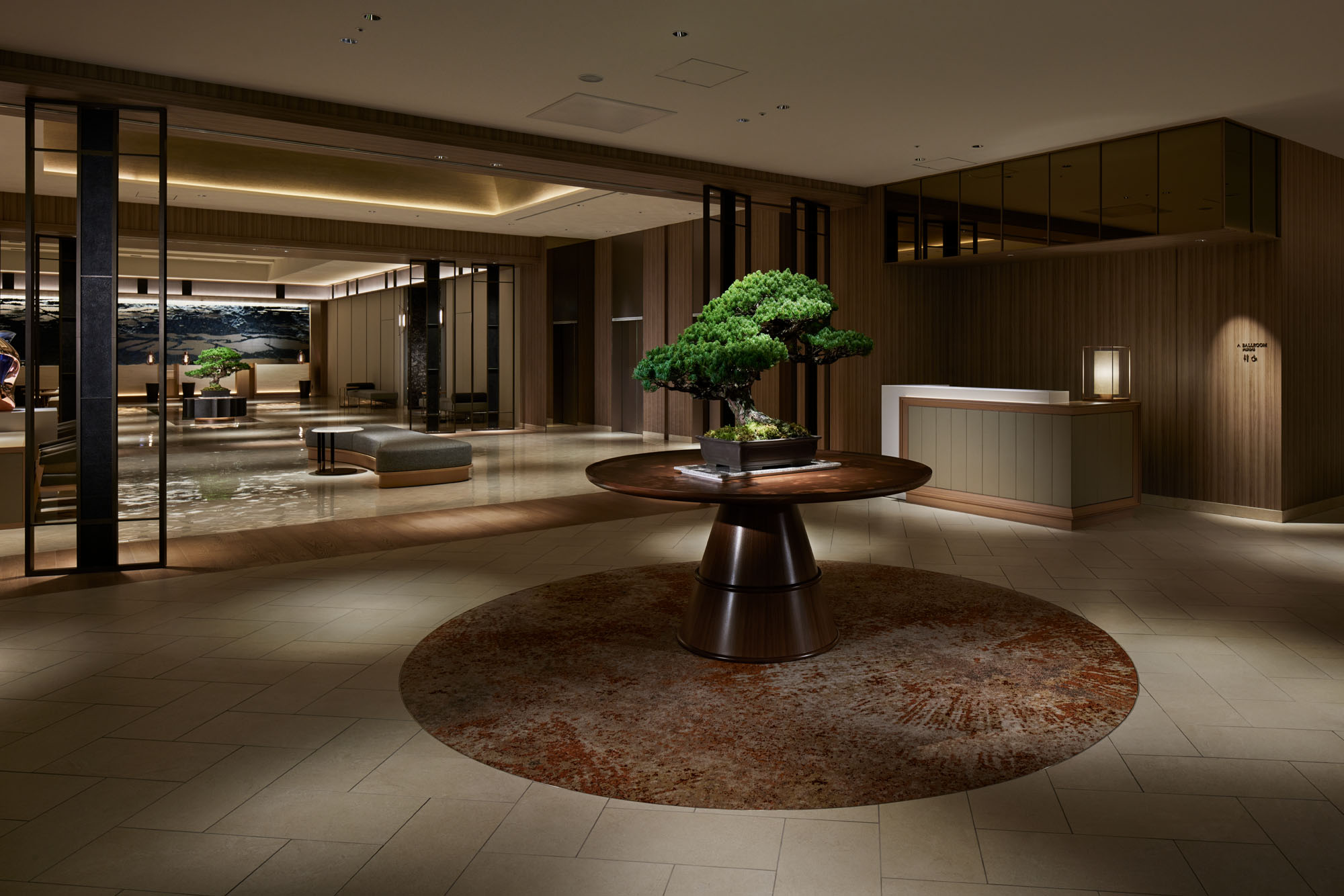 Elegant hotel lobby featuring a round wooden table with a lush bonsai centerpiece. Soft lighting and modern decor create a serene atmosphere.