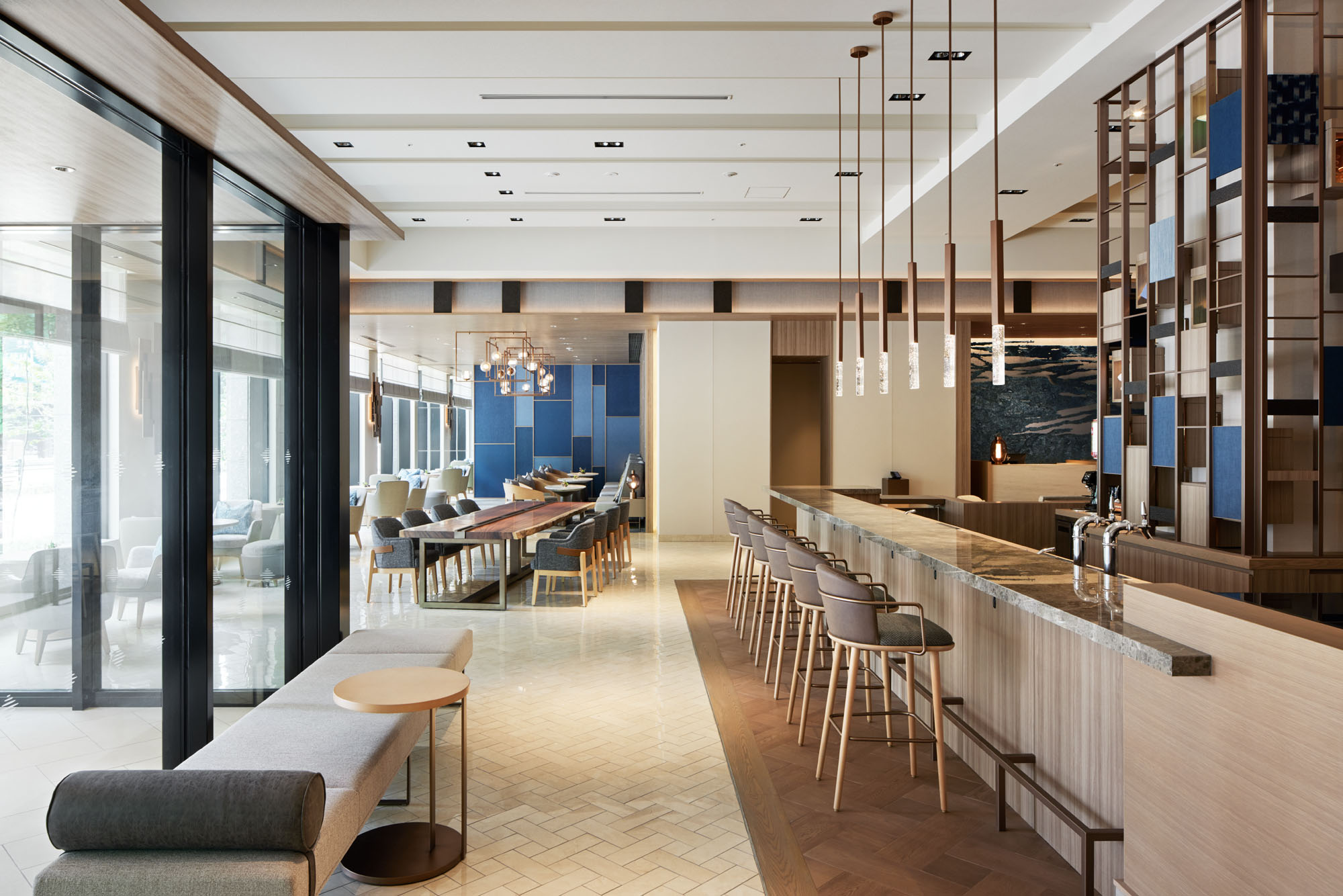 Spacious modern hotel lounge with a long bar lined with stools, wooden furniture, pendant lights, and large windows. The atmosphere is sleek and inviting.