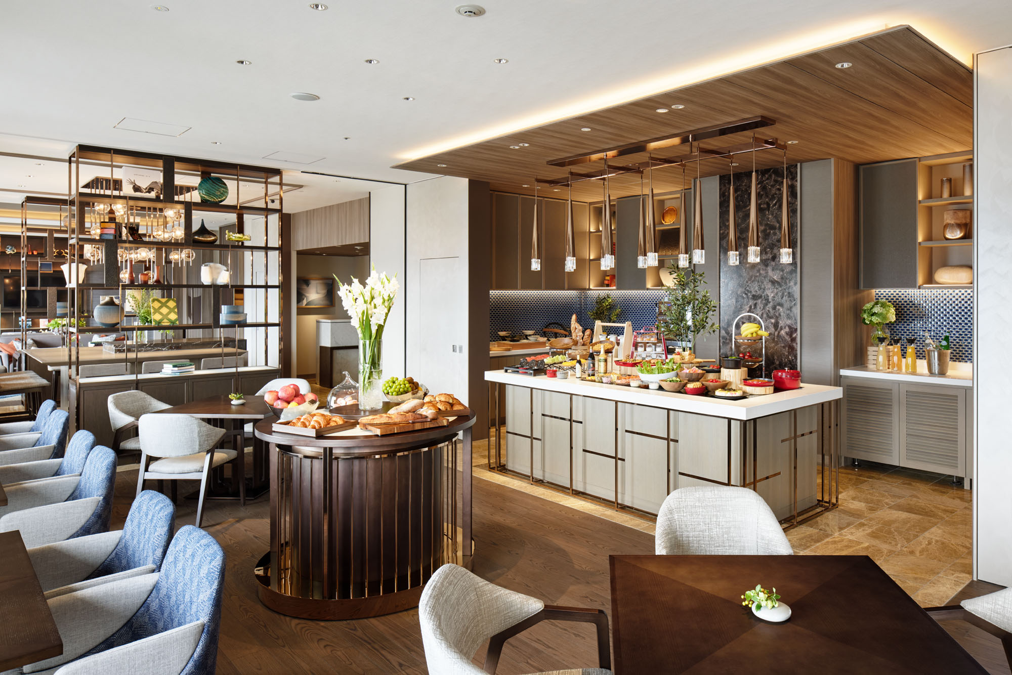 Modern hotel all day dining area featuring a kitchen with a stylish island and pendant lights. Dining chairs surround tables, and shelves display colorful fruit. Bright, inviting atmosphere.