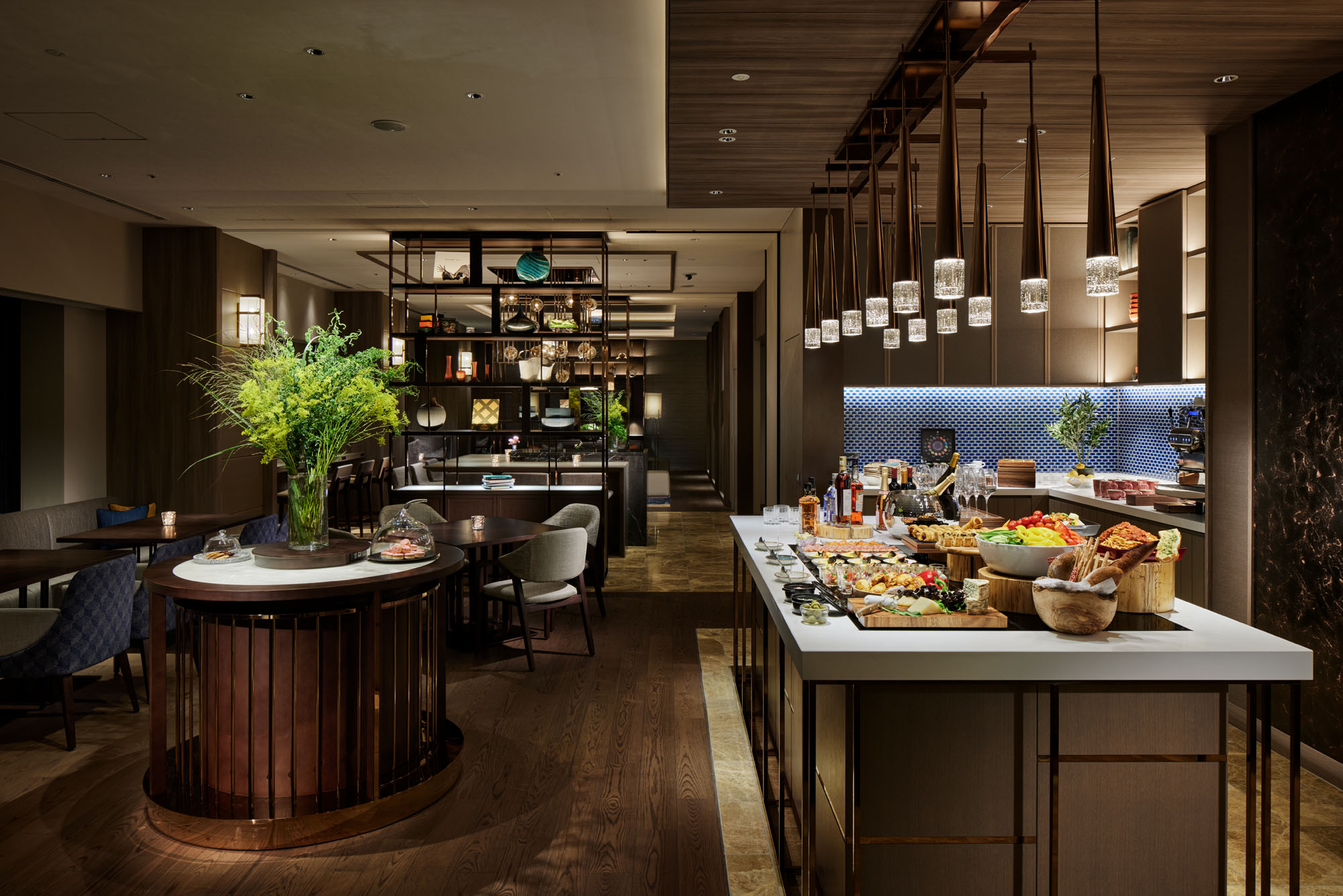 Elegant hotel dining area with a buffet table featuring sushi, fruit, and bread. Modern décor includes pendant lights, wooden accents, and lush plants.