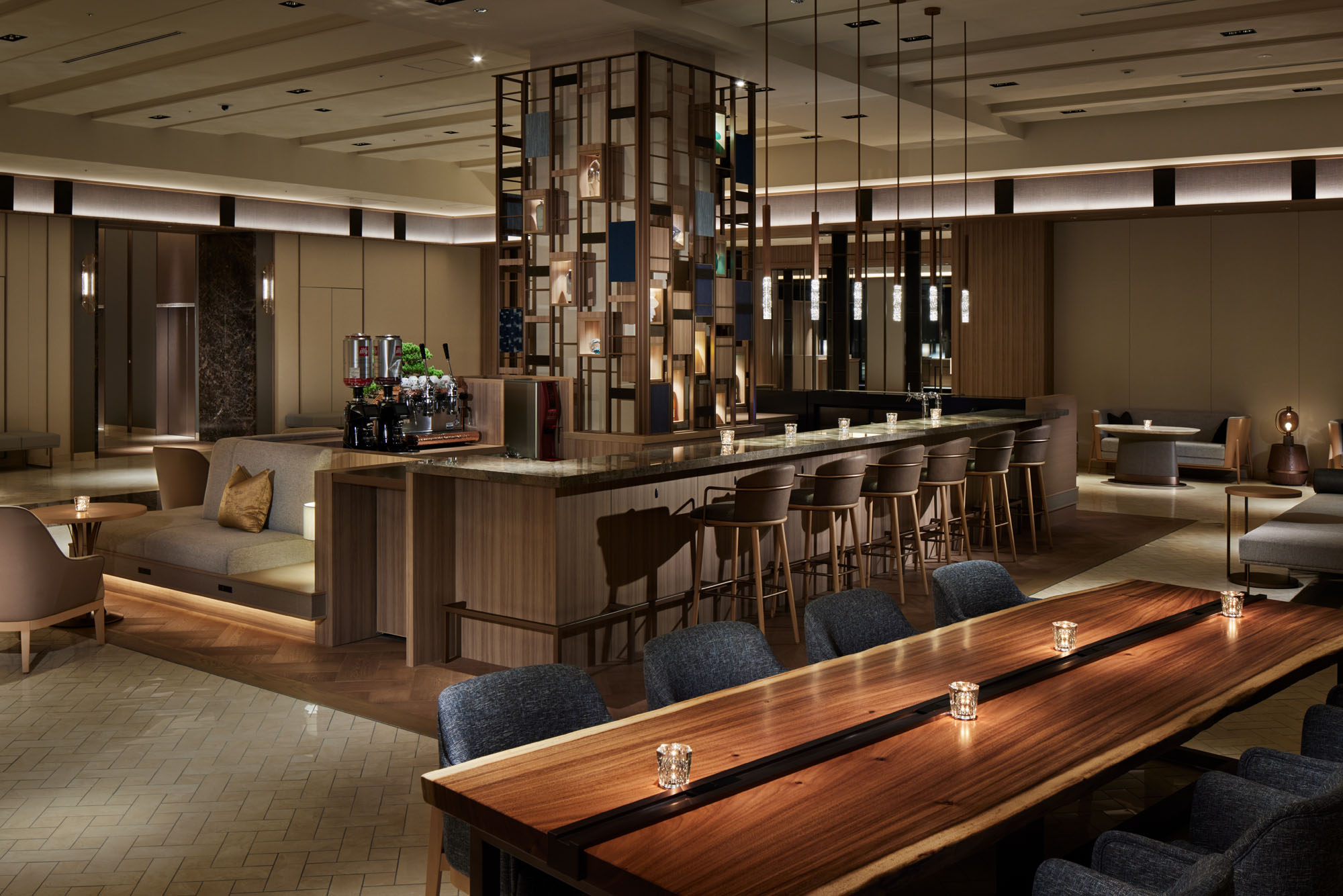 Modern hotel lounge bar with warm lighting and cozy ambiance. Features a long wooden bar with high chairs, a mosaic pillar, and a large wooden communal table.