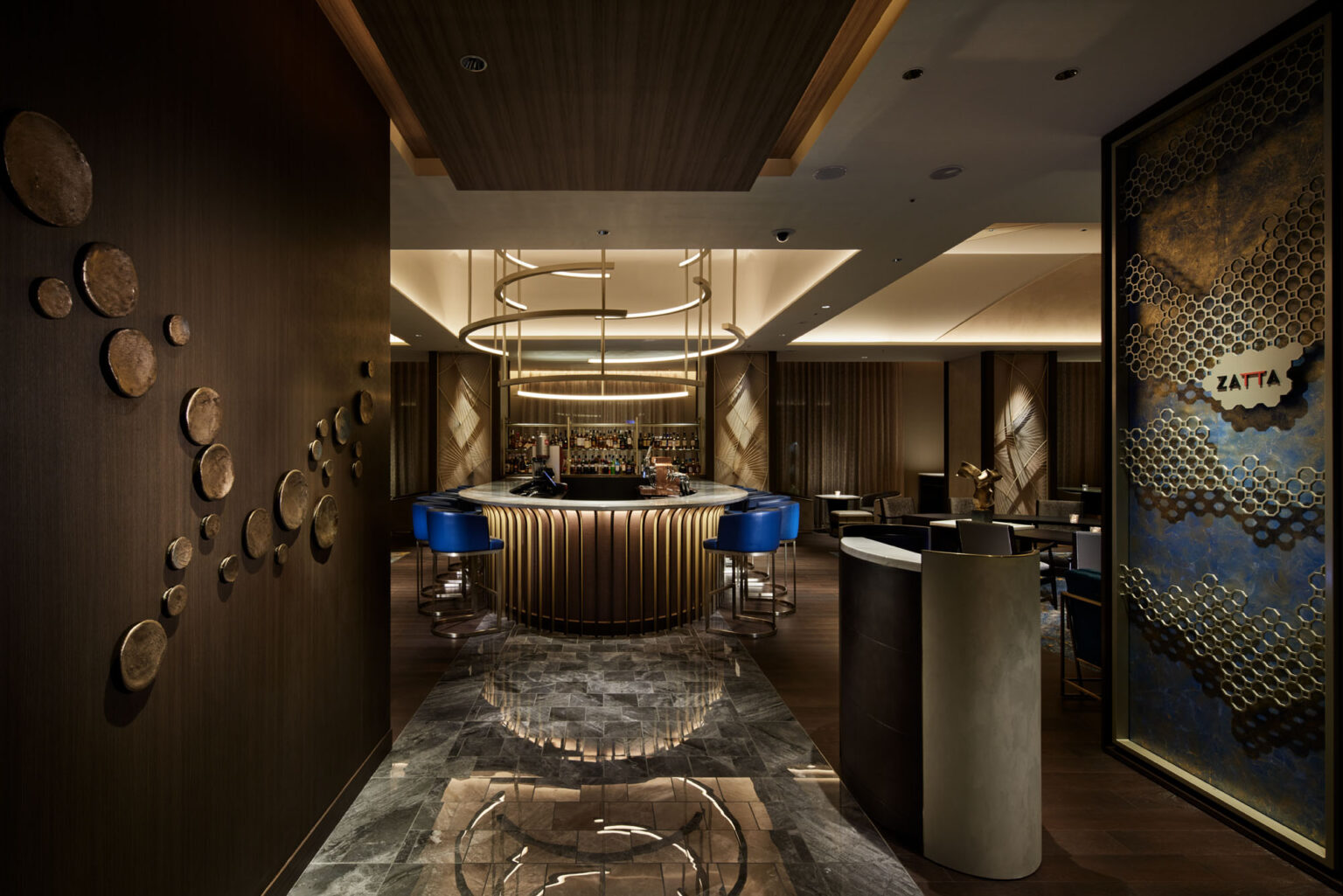 A modern, dimly-lit hotel bar features a circular counter with gold accents and blue stools. Decorative wall art and wooden elements create an elegant atmosphere.