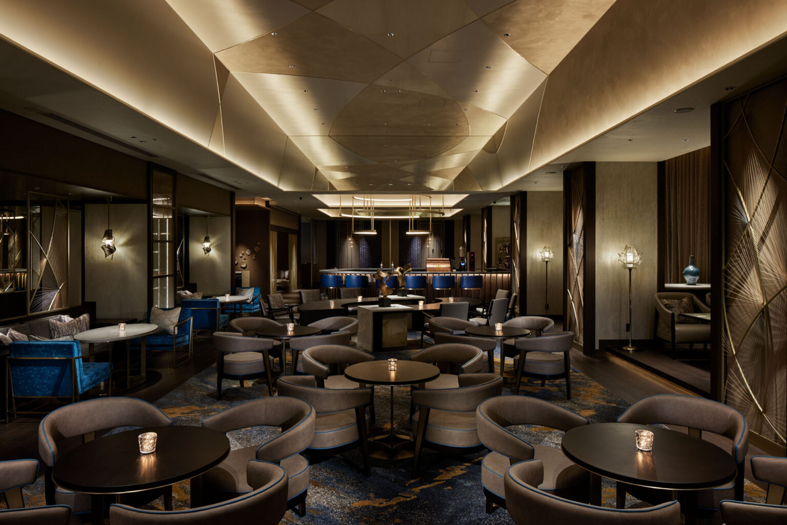 Elegant hotel lounge with dim lighting, featuring round tables, plush chairs, and a sleek bar in the background. The ambiance is cozy and sophisticated.