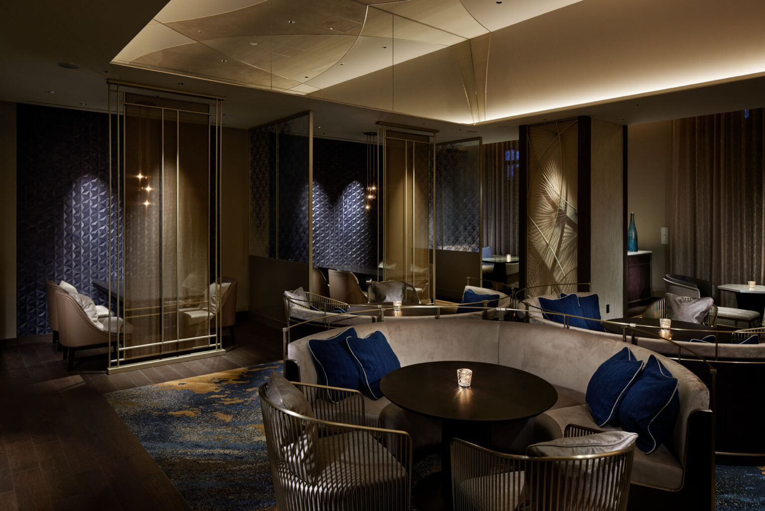 Elegant hotel lounge with soft lighting features round seating, blue cushions, and a dark table. Textured walls and subtle decor create a cozy, modern atmosphere.