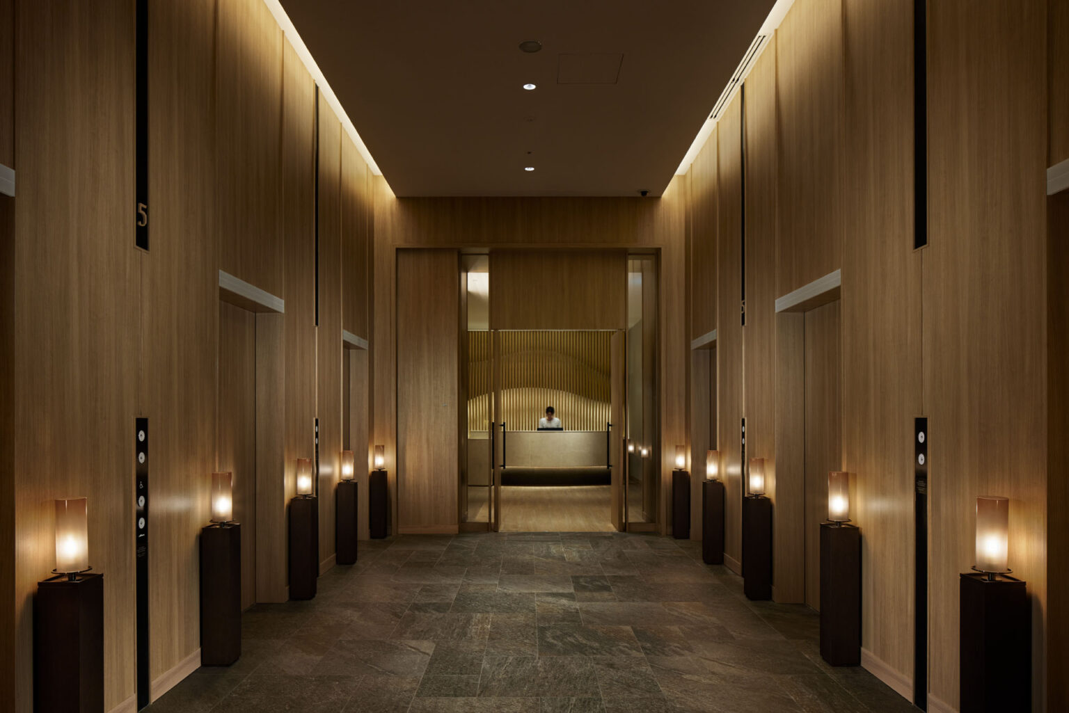 Dimly lit modern hotel lobby with wooden walls and soft lighting. A distant reception desk, flanked by elevators, creates a calm, welcoming ambiance.
