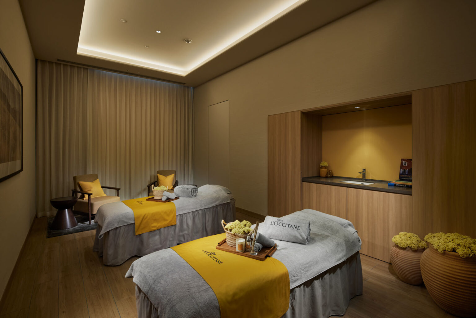 Calm hotel spa treatment room with two massage beds covered in gray and yellow linens. Soft lighting, wooden decor, and potted flowers create a serene atmosphere.